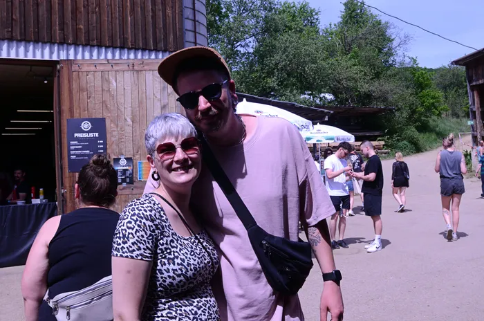 Ein Paar lächelt in die Kamera, vor ihnen ein Holzgebäude und im Hintergrund Menschen auf einem Festivalgelände.