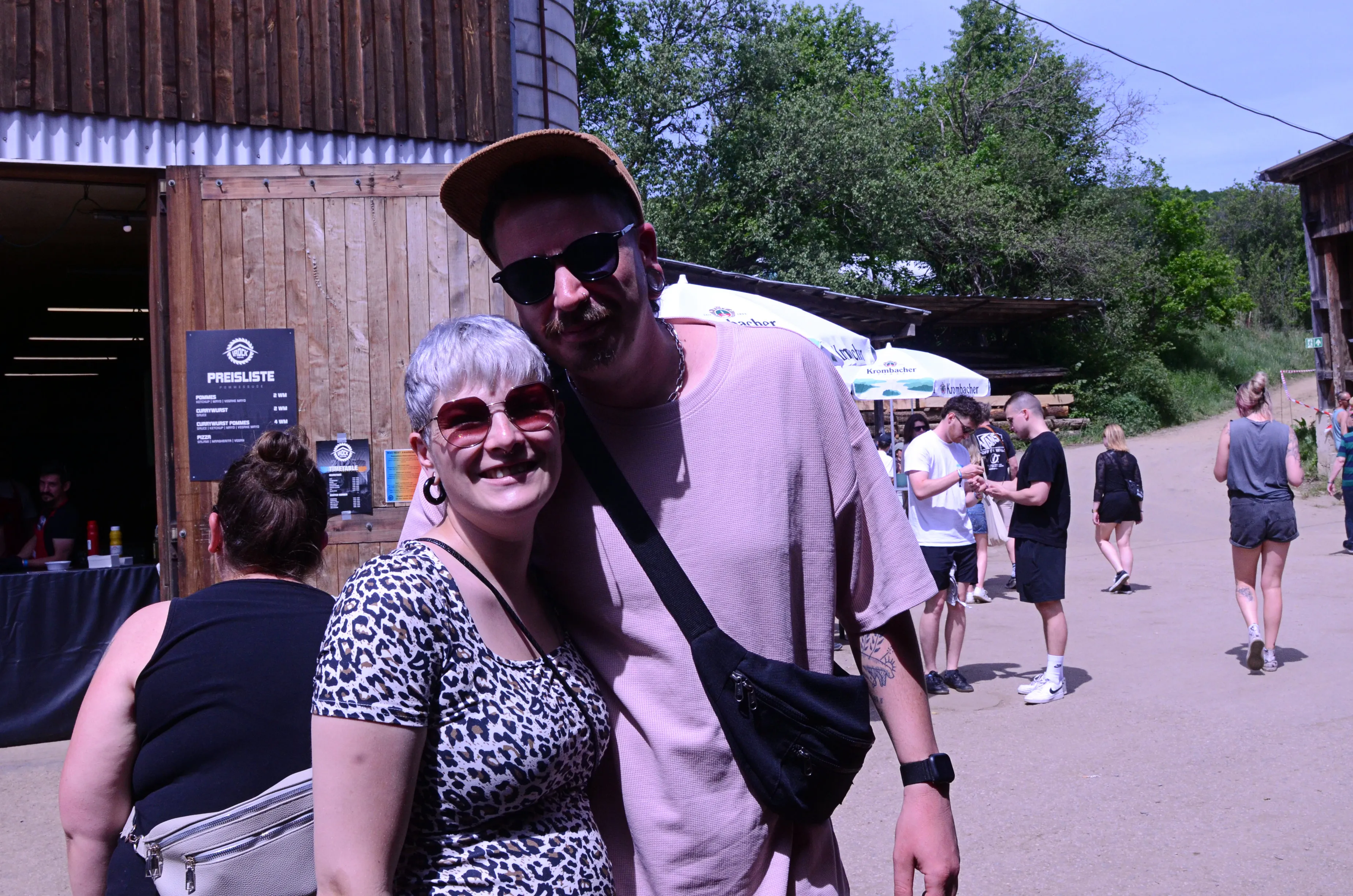 Ein Paar lächelt in die Kamera, vor ihnen ein Holzgebäude und im Hintergrund Menschen auf einem Festivalgelände.