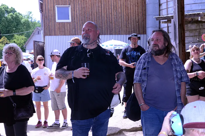 Zuschauer stehen auf einem Festivalgelände vor einer Scheune, ein Mann mit Tattoos hält ein Bierglas in der Hand.