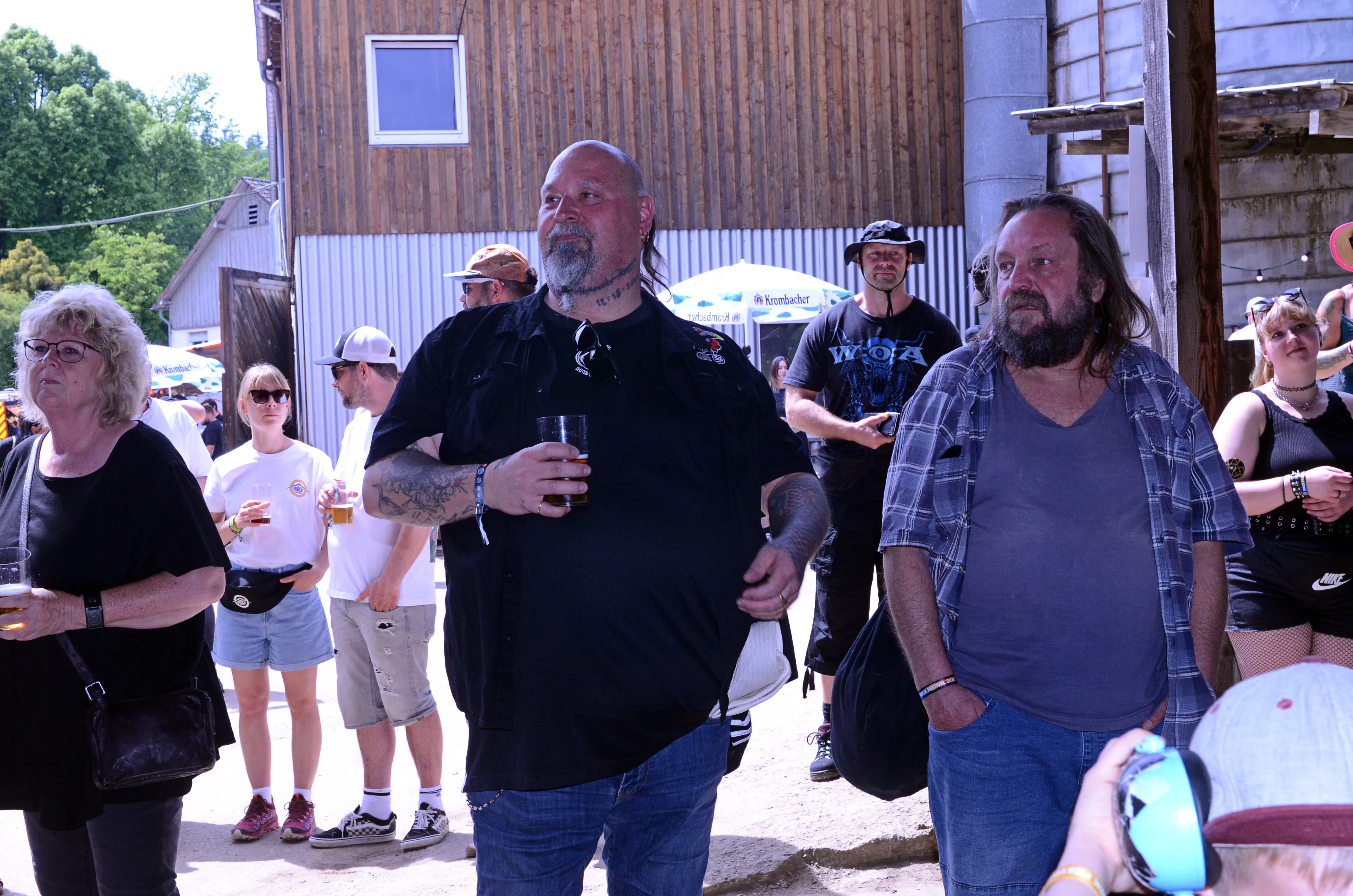 Zuschauer stehen auf einem Festivalgelände vor einer Scheune, ein Mann mit Tattoos hält ein Bierglas in der Hand.