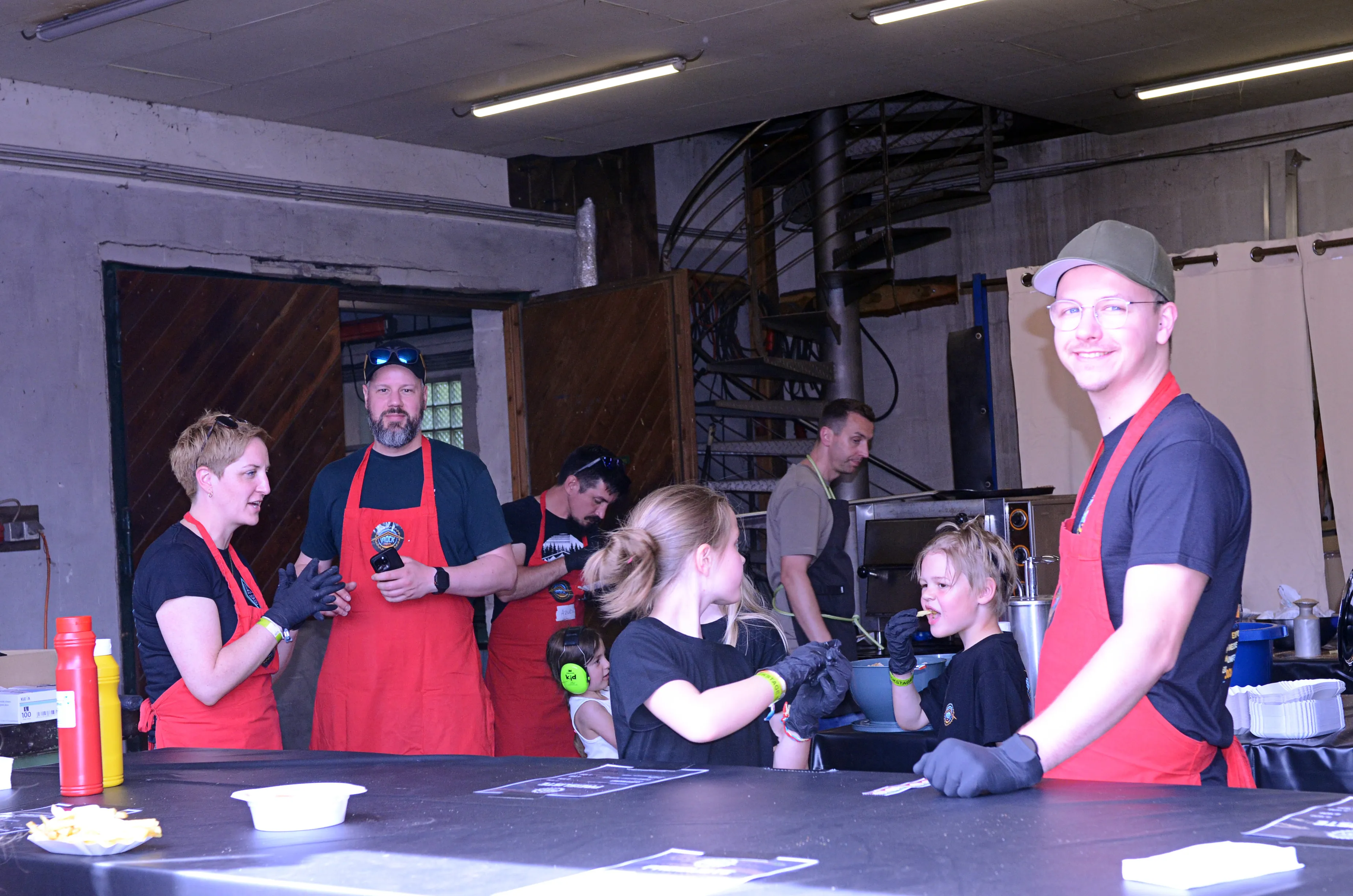 Mehrere Personen in roten Schürzen bereiten Speisen an einem mobilen Imbissstand auf dem I-Rock Festival zu.