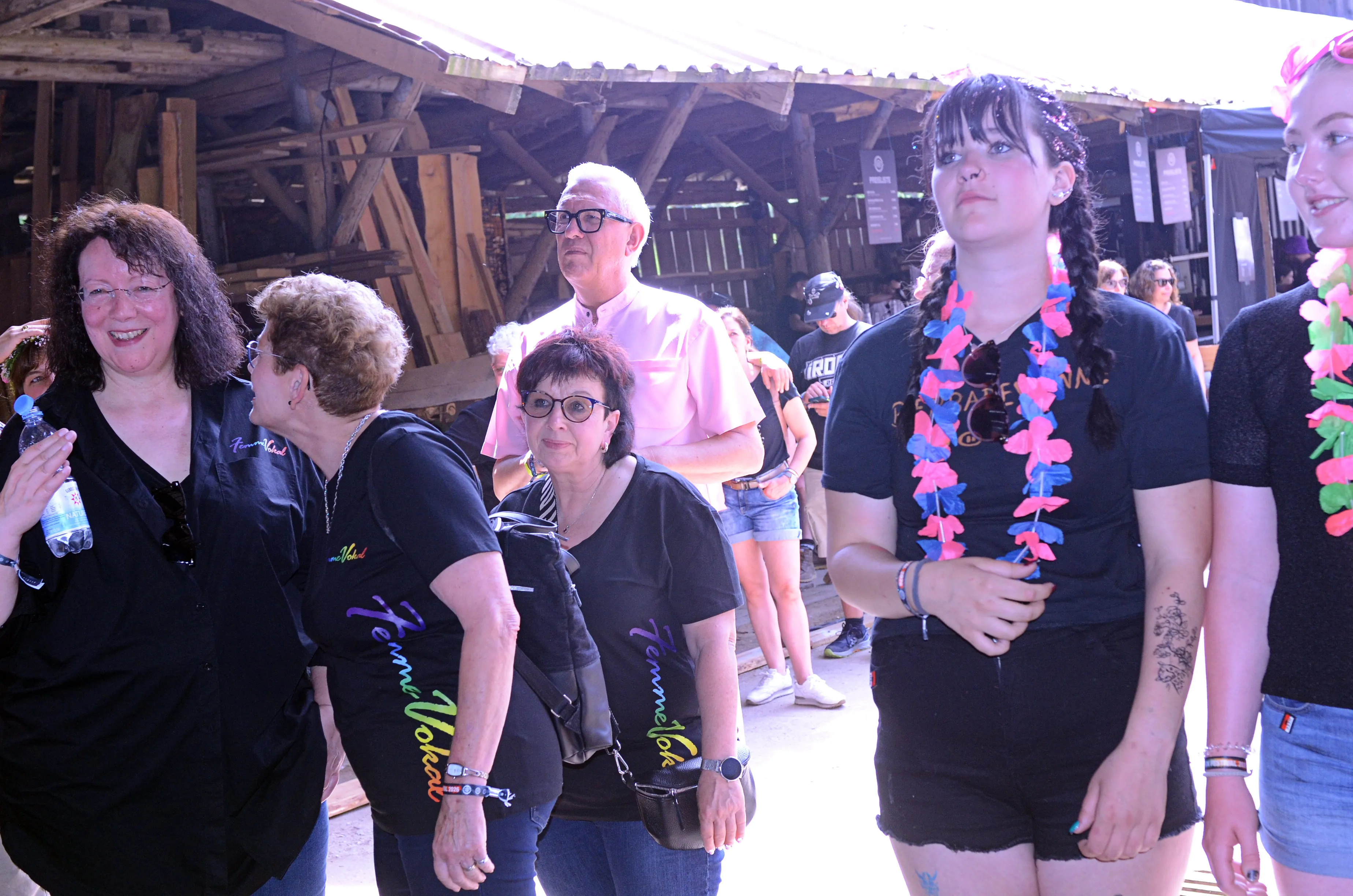 Mehrere Menschen gehen auf einem Festivalgelände vorbei, einige tragen schwarze T-Shirts mit dem Aufdruck „Femme Vokal“ und Blumenketten.