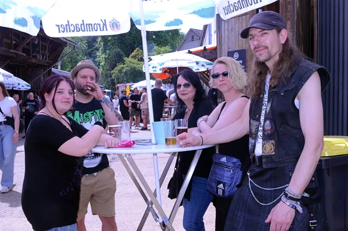 Fünf Personen stehen um einen kleinen Tisch auf dem Gelände des Immecke Rock Festivals. Sie halten Biergläser und unterhalten sich.