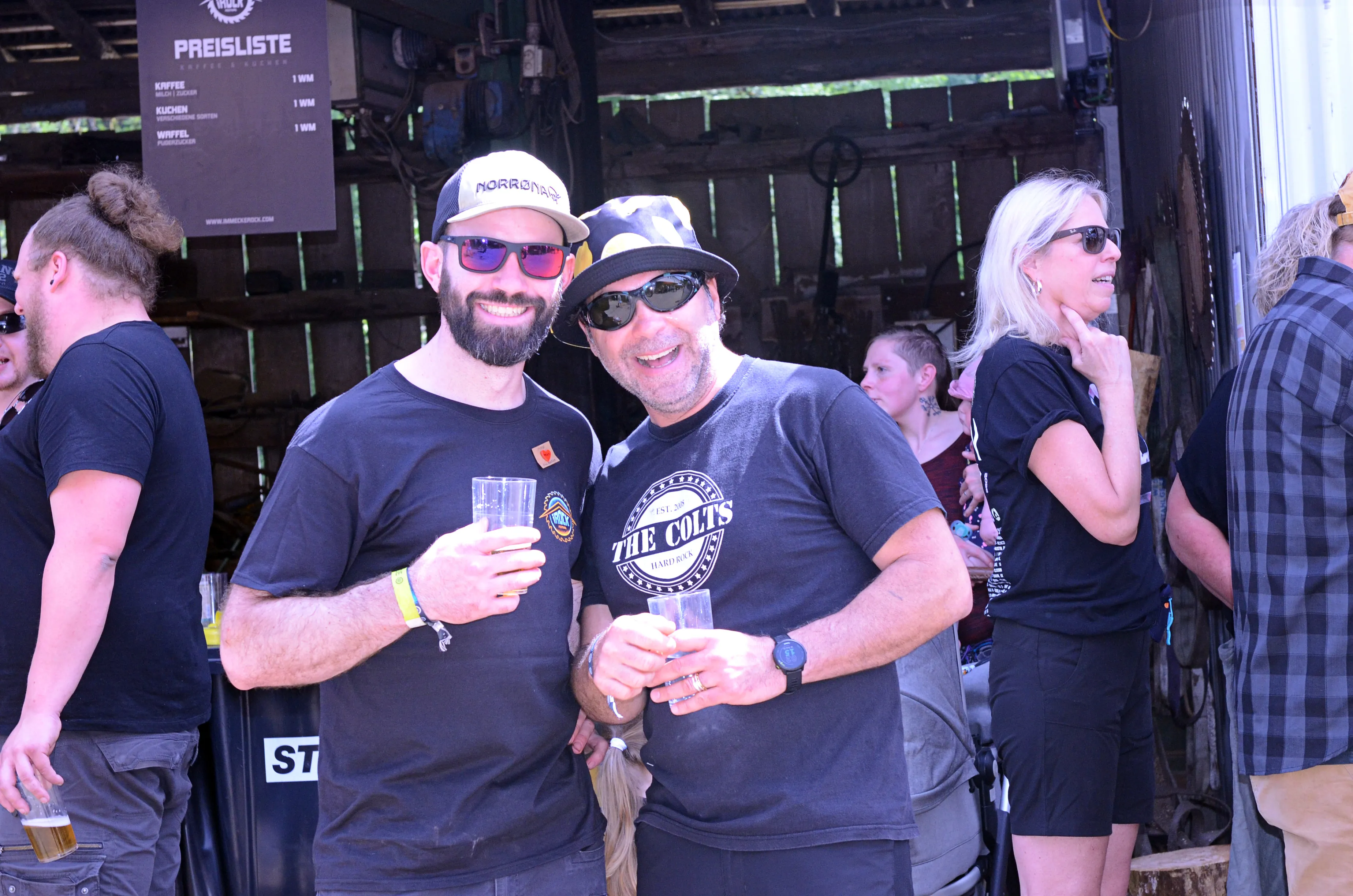 Zwei Männer mit Sonnenbrillen und ein Bier in der Hand stehen vor einer Menschenmenge auf dem Immecke Rock Festival.