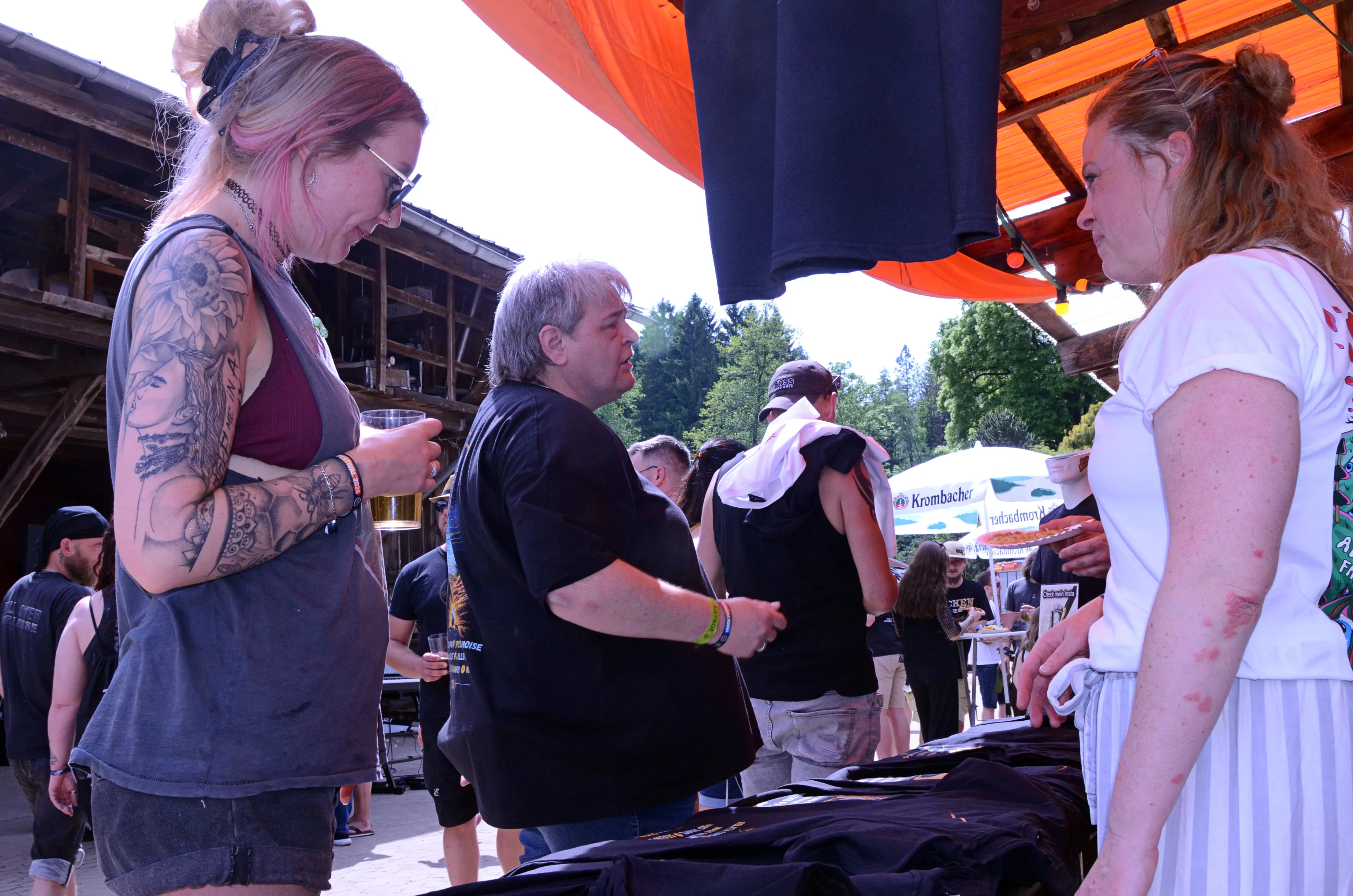 Zwei Frauen und ein Mann stehen an einem Verkaufsstand mit T-Shirts auf dem Immecke Rock Festival in Plettenberg.