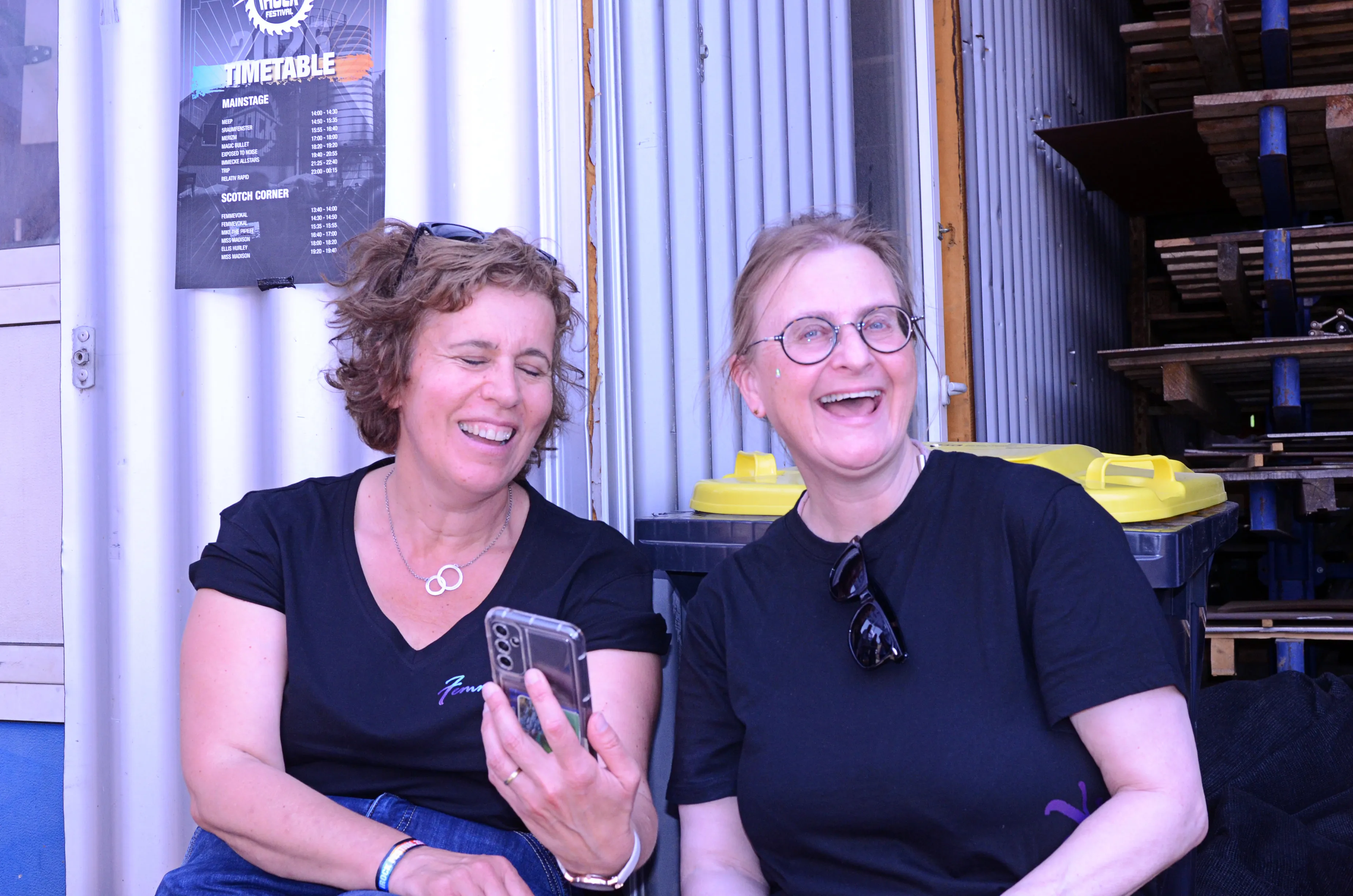 Zwei lachende Frauen sitzen nebeneinander und schauen auf ein Smartphone. Im Hintergrund ist ein Festivalplan zu sehen.