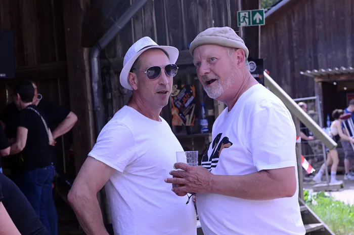 Zwei lächelnde Männer im mittleren Alter stehen eng beieinander vor einer Holzwand, einer hält ein Glas in der Hand. Im Hintergrund sind weitere Festivalbesucher zu sehen.