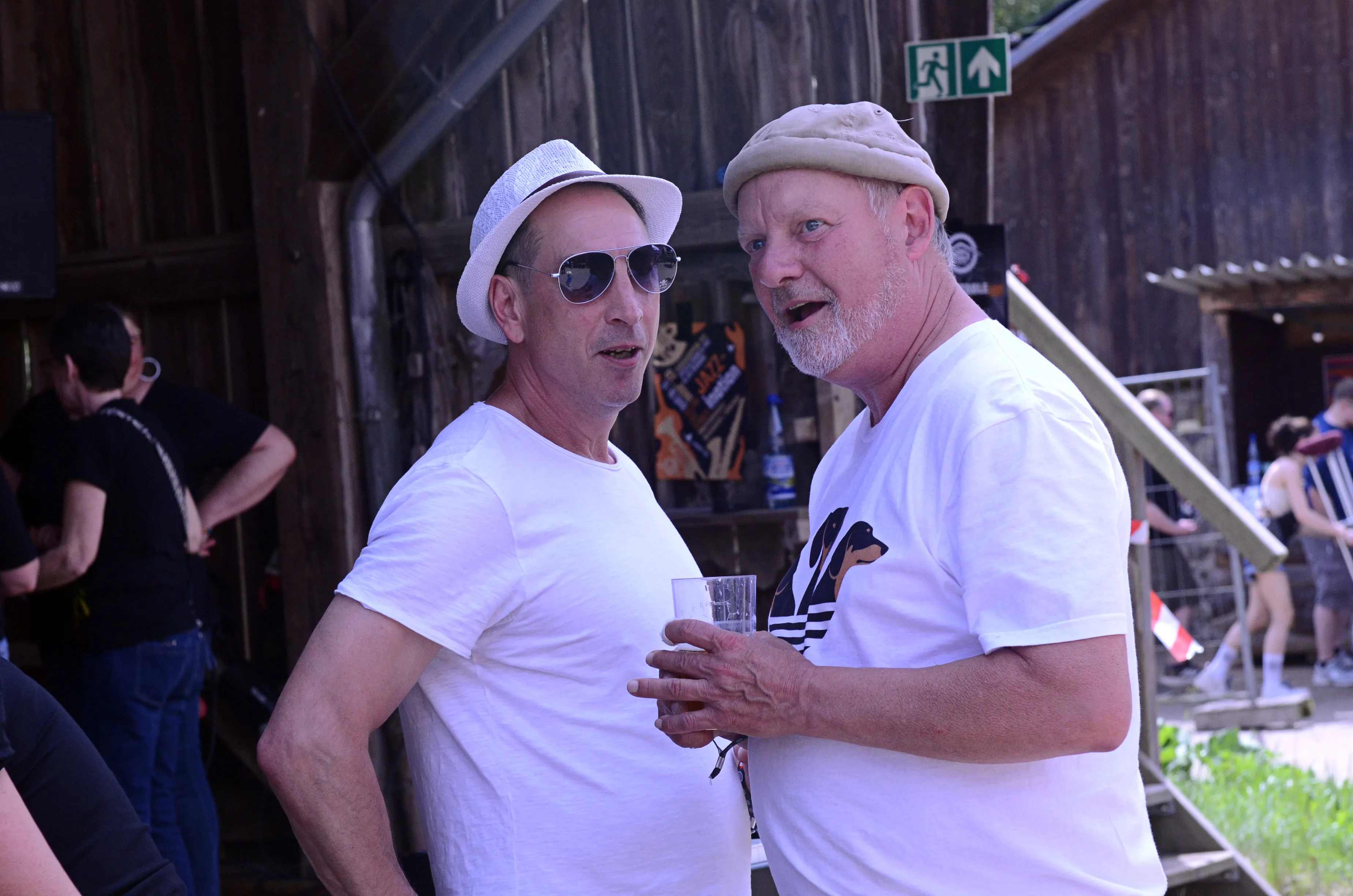 Zwei lächelnde Männer im mittleren Alter stehen eng beieinander vor einer Holzwand, einer hält ein Glas in der Hand. Im Hintergrund sind weitere Festivalbesucher zu sehen.