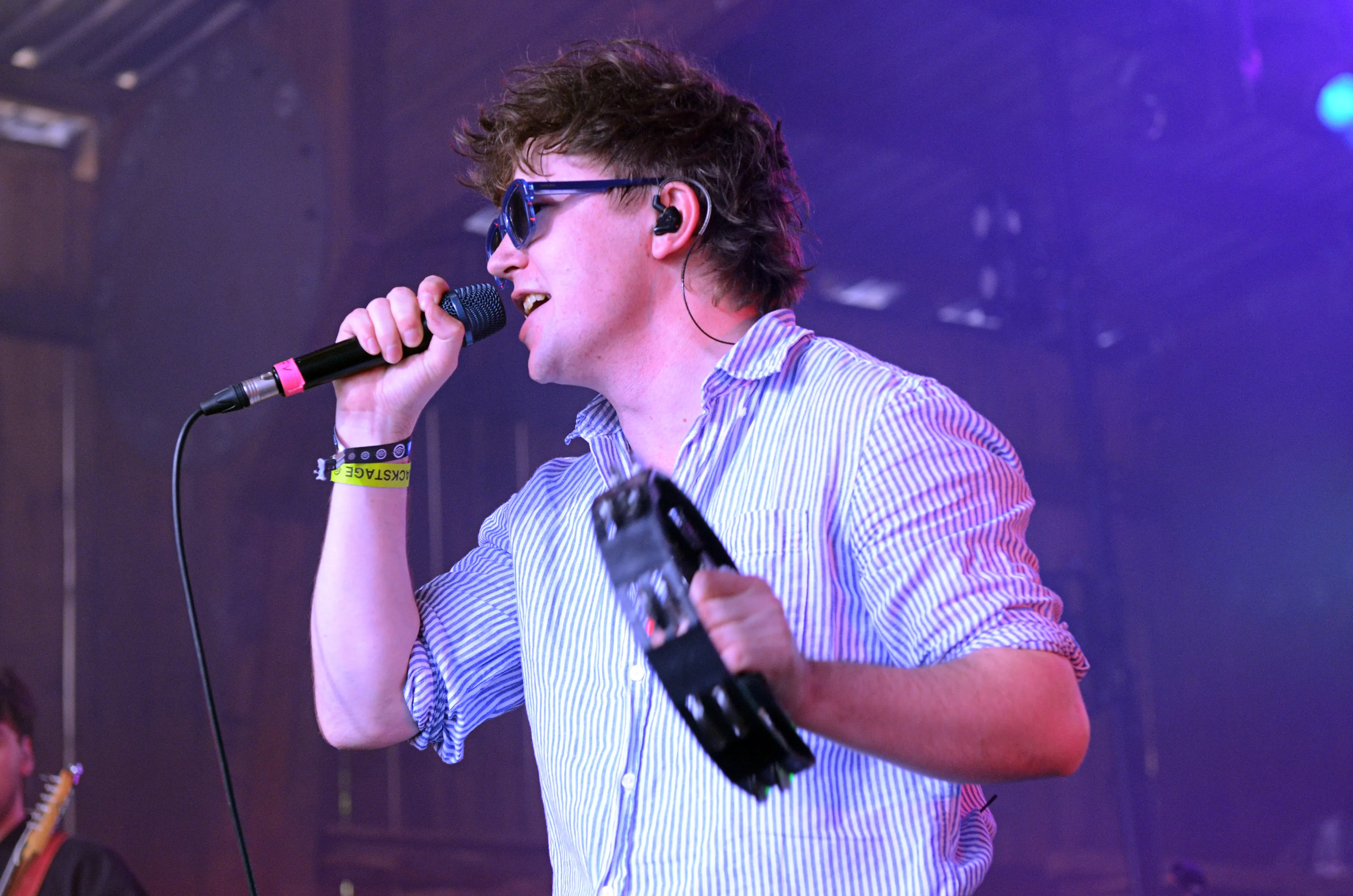 Sänger mit dunklen lockigen Haaren und Sonnenbrille singt in ein Mikrofon während eines Konzerts auf der Bühne des Immecke Rock Festivals.