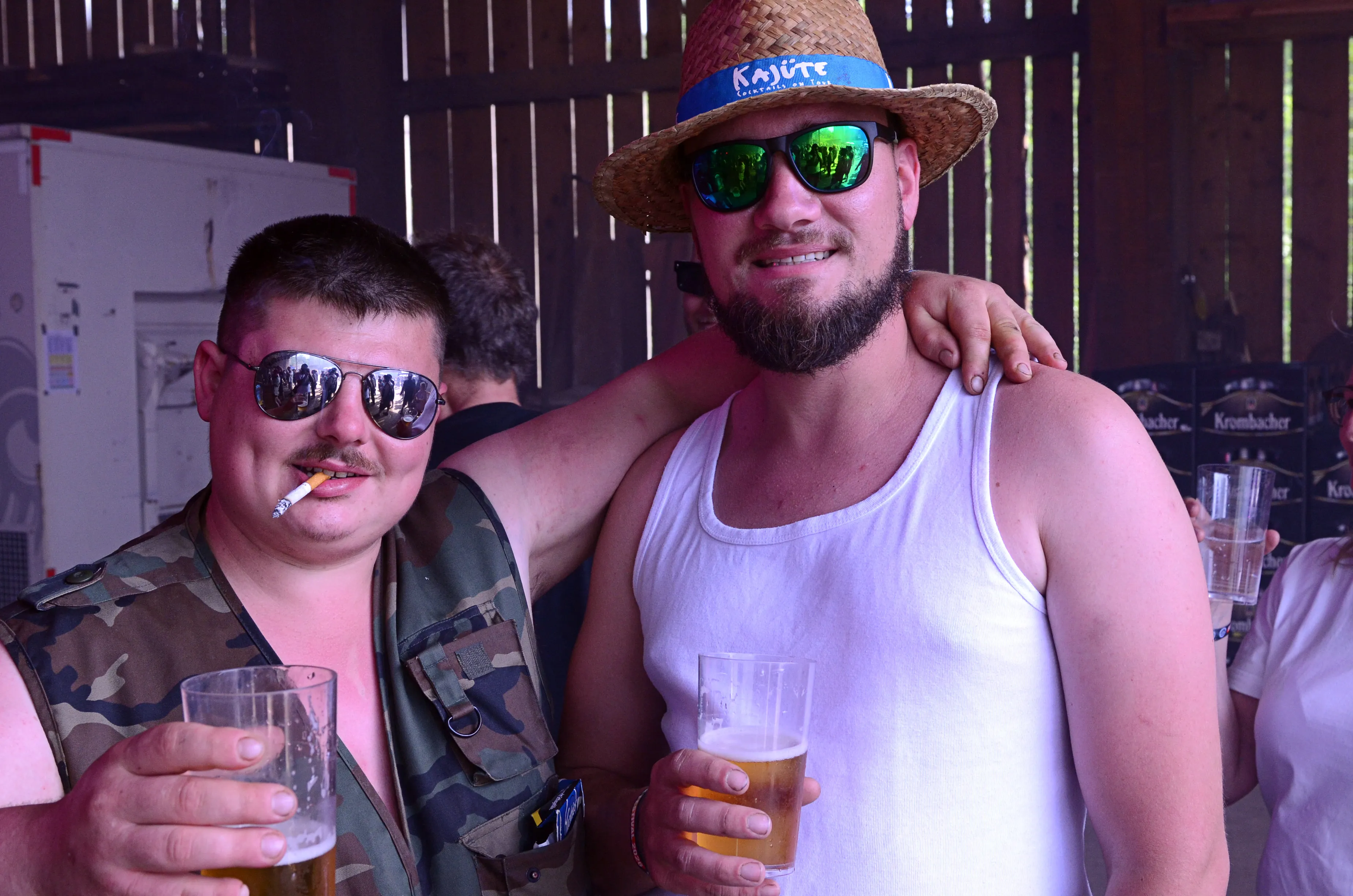 Zwei Männer mit Sonnenbrillen und Biergläsern posieren eng umschlungen. Im Hintergrund sind weitere Festivalbesucher und ein Getränkestand zu sehen.