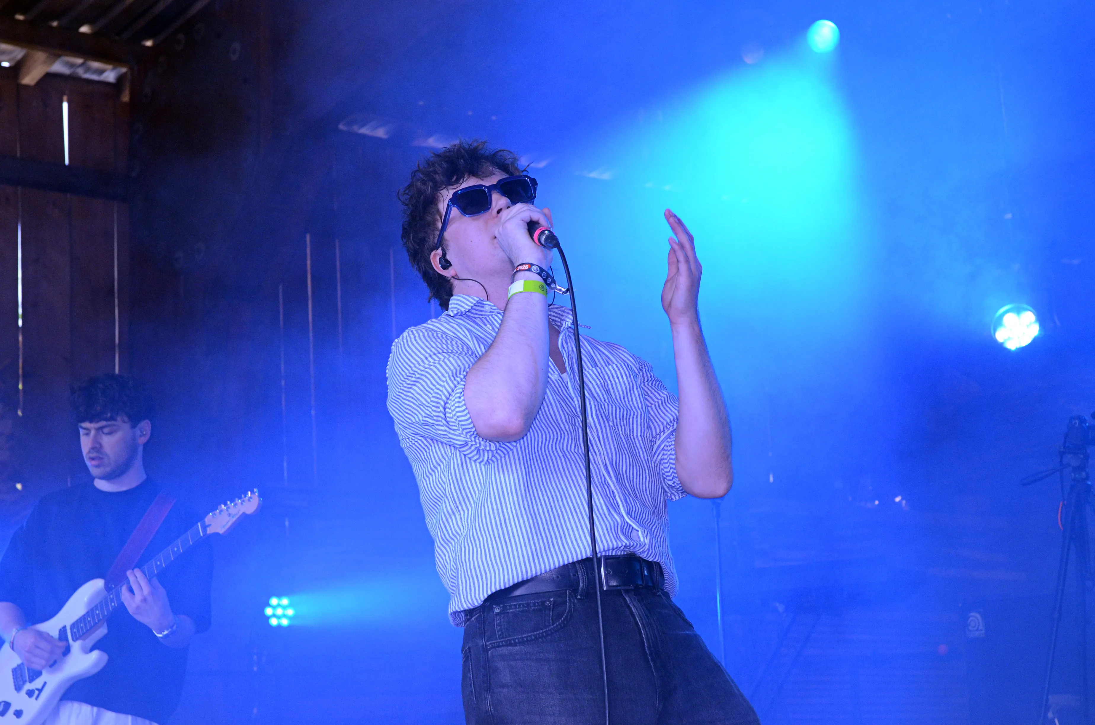 Sänger mit Sonnenbrille und offenem Hemd singt in ein Mikrofon auf der Bühne des Immecke Rock Festivals. Im Hintergrund ein Gitarrist.