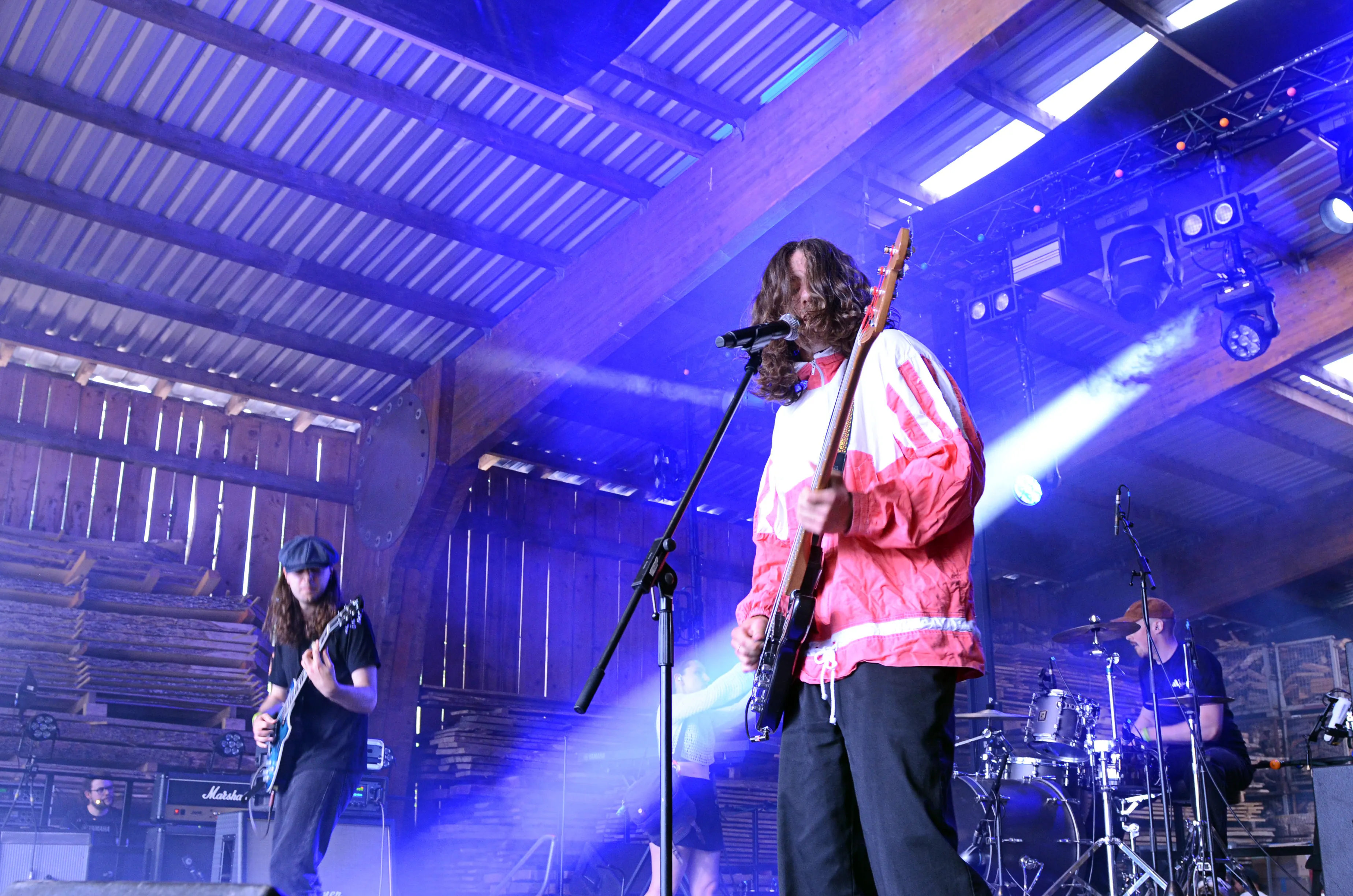 Eine Band spielt auf einer Bühne in einer großen Scheune. Der Sänger mit langen Haaren und roter Jacke steht am Mikrofon, während ein Gitarrist links im Bild spielt und ein Schlagzeuger rechts zu sehen ist.