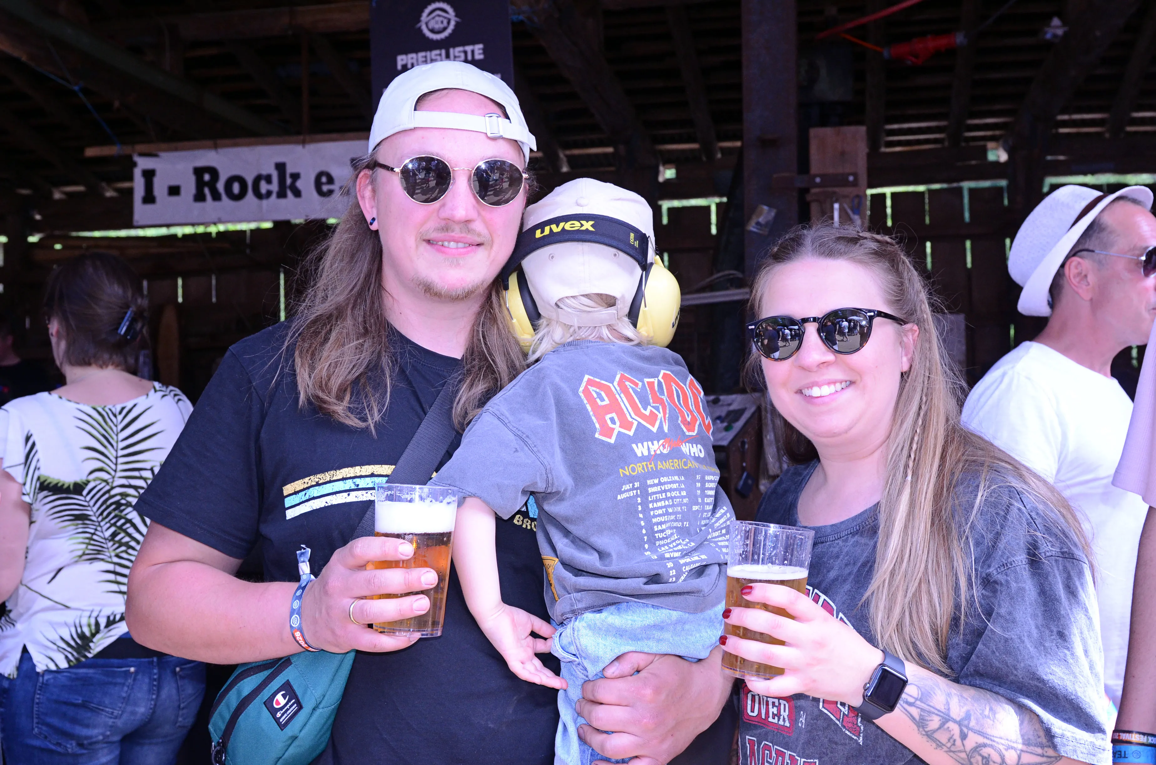 Ein Mann mit langen Haaren und eine Frau mit einem kleinen Kind im AC/DC-T-Shirt stehen auf dem I-Rock Festival in Plettenberg und halten jeweils ein Bierglas in der Hand.