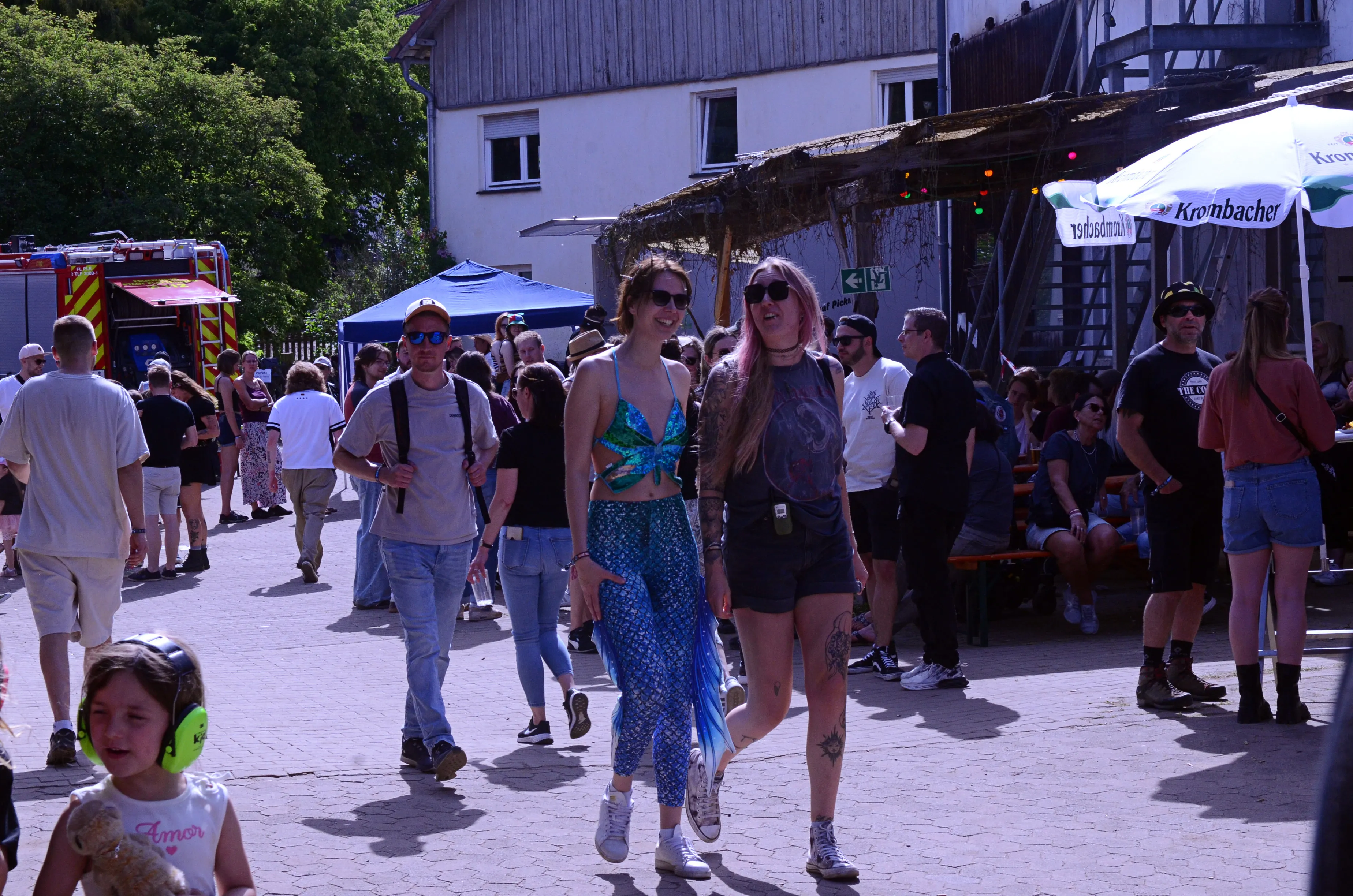 Menschenmengen bewegen sich auf einem Festivalgelände mit Bühnen und Essensständen. Im Hintergrund ein rotes Feuerwehrfahrzeug.