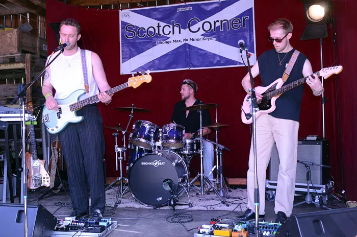 Band spielt auf einer Bühne vor rotem Vorhang. Im Hintergrund ein Banner mit dem Aufdruck „Scotch Corner“ und dem Slogan „3 Songs Max, No Minor Keys“.