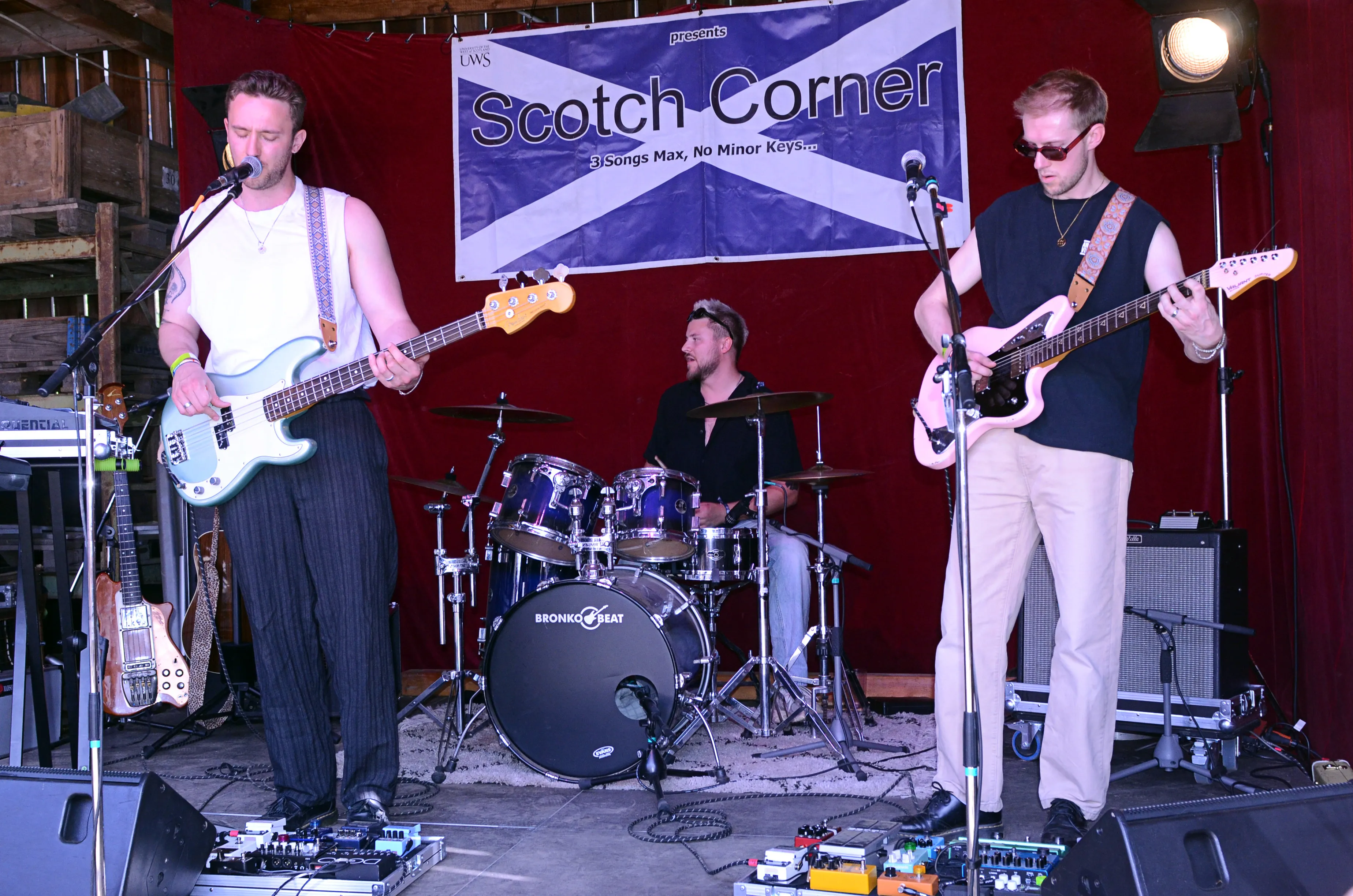 Band spielt auf einer Bühne vor rotem Vorhang. Im Hintergrund ein Banner mit dem Aufdruck „Scotch Corner“ und dem Slogan „3 Songs Max, No Minor Keys“.