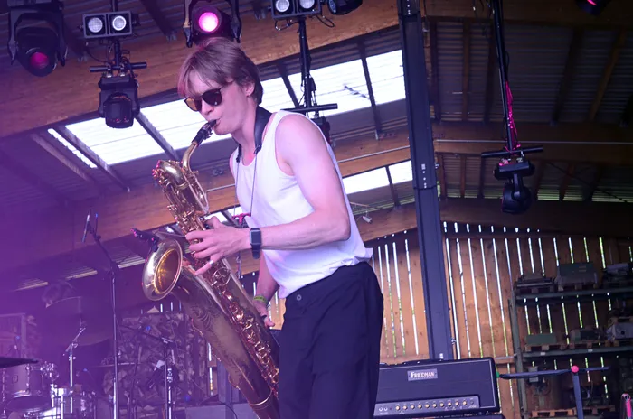 Ein Mann mit Sonnenbrille spielt auf der Bühne Saxophon. Im Hintergrund sind Bühnenlichter und ein Drumset zu sehen.
