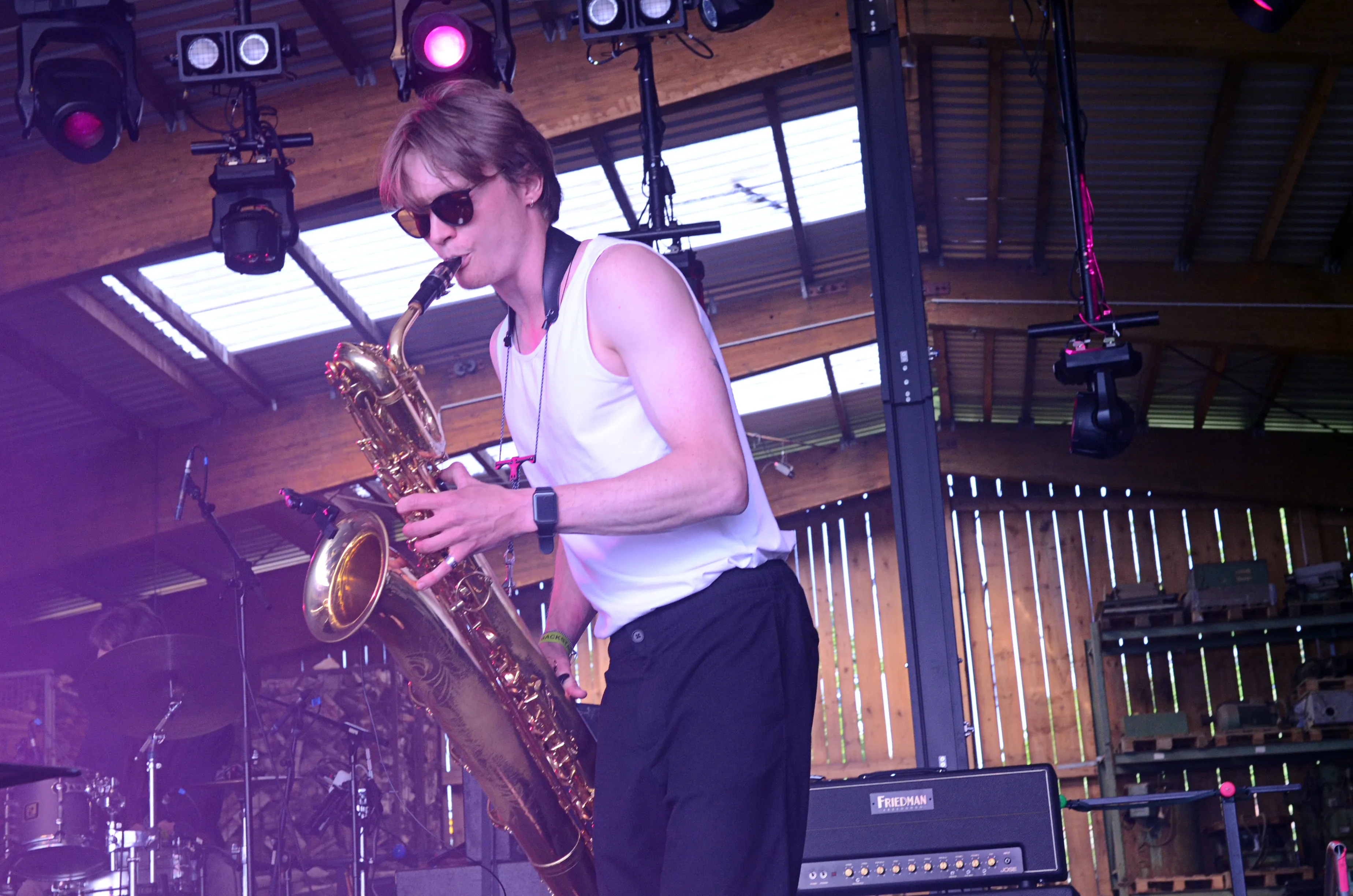 Ein Mann mit Sonnenbrille spielt auf der Bühne Saxophon. Im Hintergrund sind Bühnenlichter und ein Drumset zu sehen.