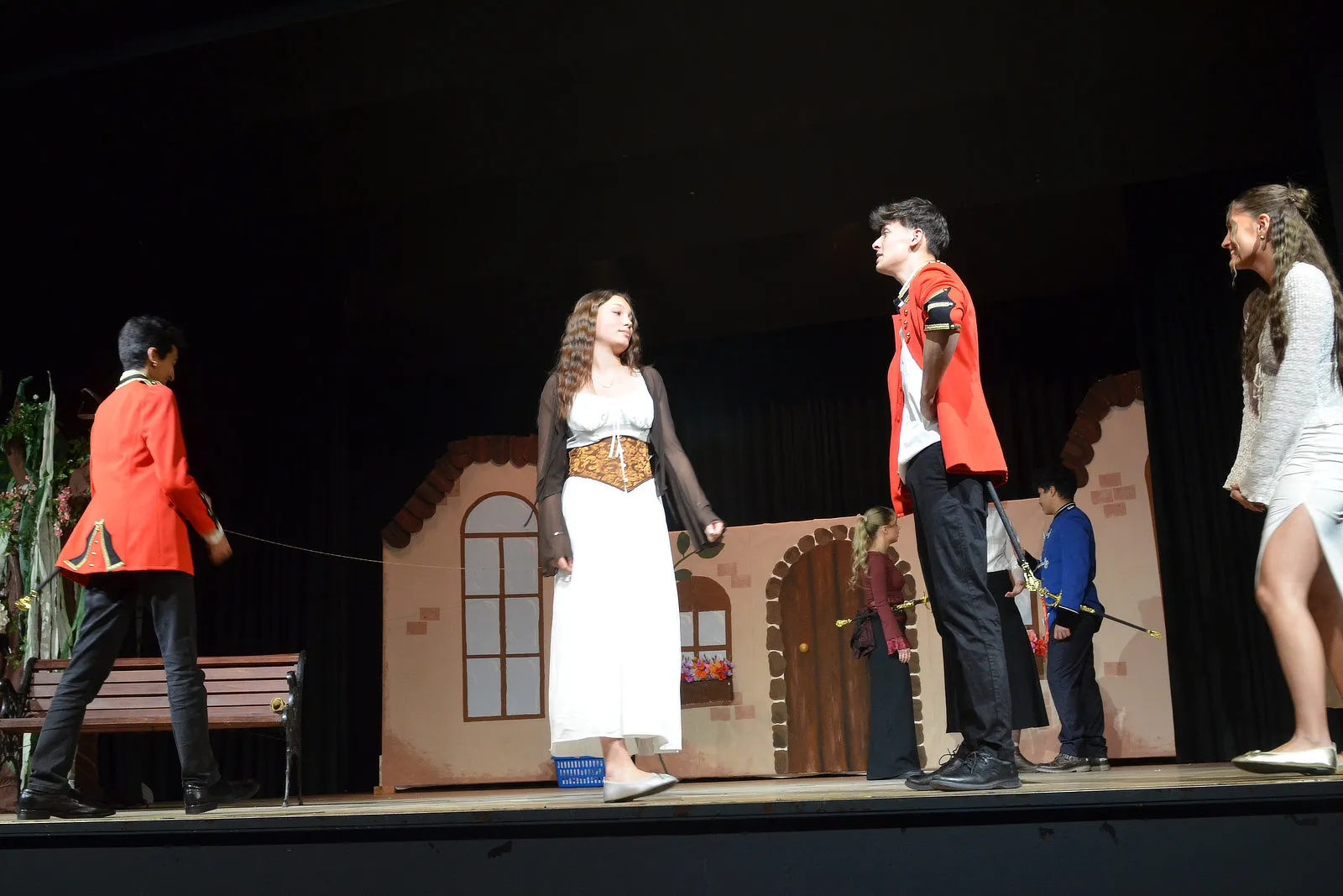 Szene aus der Theateraufführung „Viel Lärm um nichts“ mit Schülerinnen und Schülern in Kostümen vor einer Kulisse, die ein Haus darstellt.