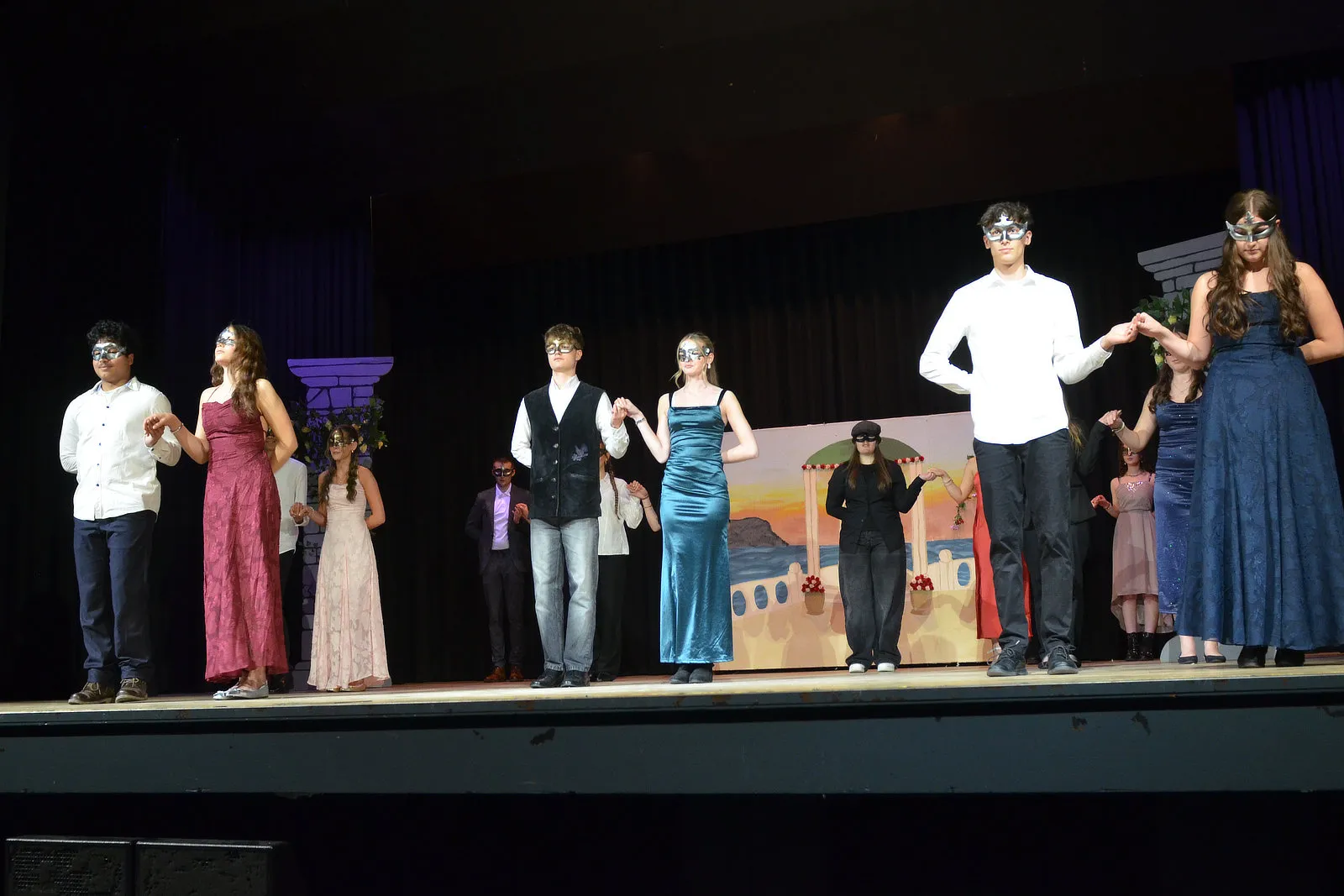 Ensemble aus Schülerinnen und Schülern in Kostümen auf einer Bühne, einige tragen Masken, vor Bühnenbildern mit Säulen und einer Burg. Sie spielen das Theaterstück „Viel Lärm um nichts“.