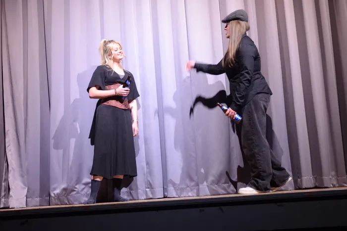 Zwei Darsteller auf einer Bühne führen eine Szene aus Shakespeares „Viel Lärm um nichts“ auf. Die Frau links hält ein Mikrofon, während der Mann rechts eine Geste macht und eine Wasserflasche in der Hand hält.