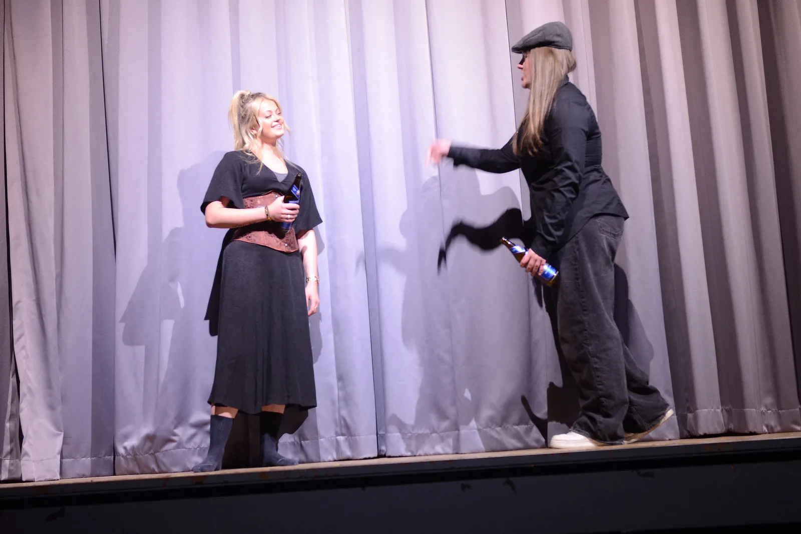 Zwei Darsteller auf einer Bühne führen eine Szene aus Shakespeares „Viel Lärm um nichts“ auf. Die Frau links hält ein Mikrofon, während der Mann rechts eine Geste macht und eine Wasserflasche in der Hand hält.