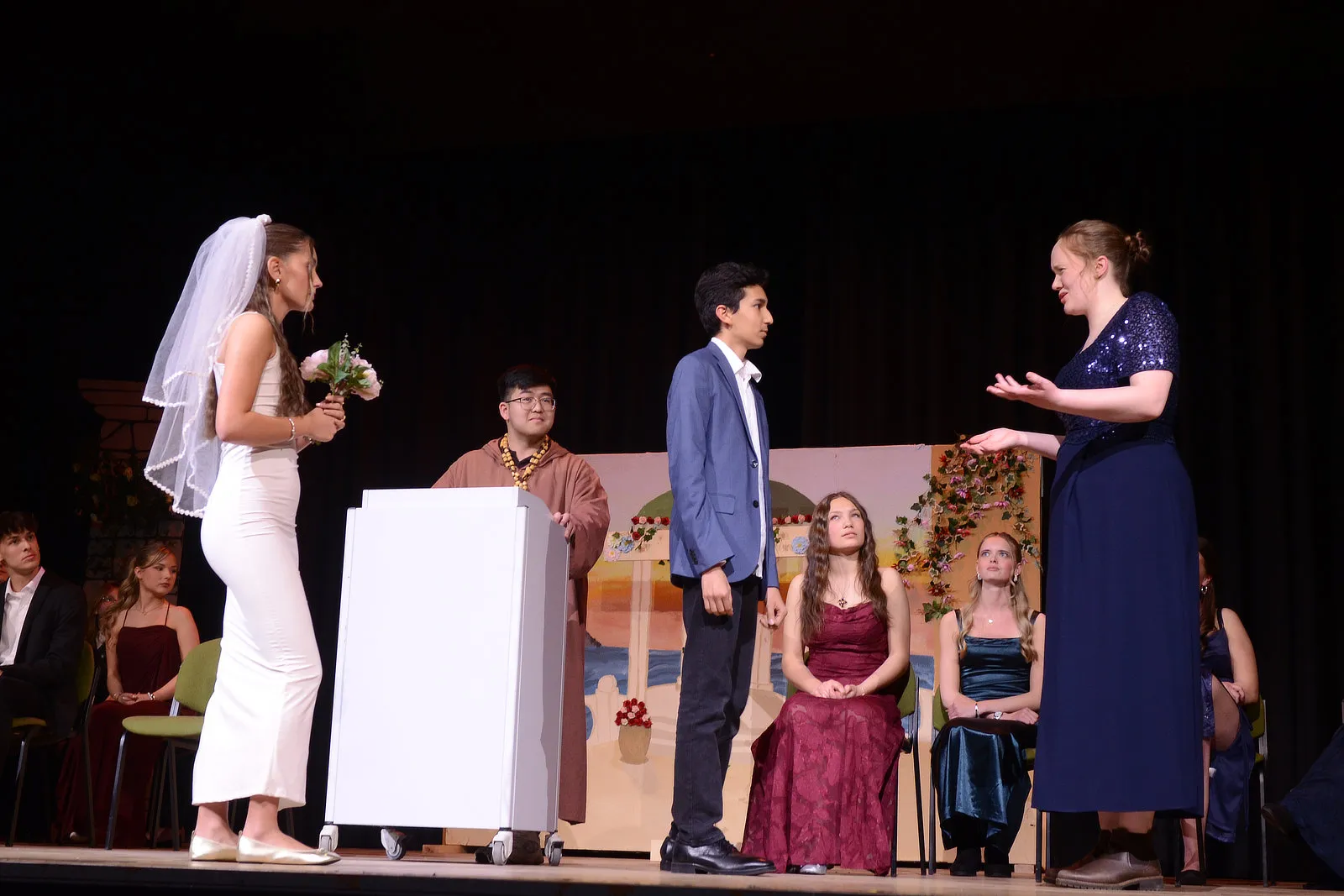 Szene aus Shakespeares „Viel Lärm um nichts“ mit einer Darstellerin im Brautkleid, einem Mann im Anzug und weiteren Ensemblemitgliedern auf einer Bühne.
