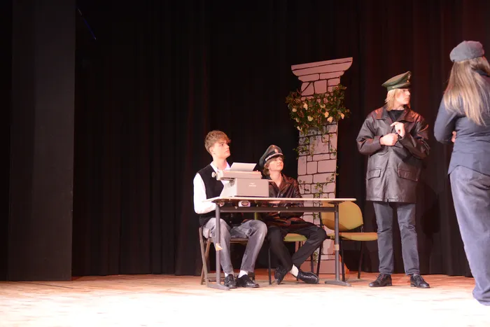 Szene aus der Theateraufführung „Viel Lärm um nichts“ mit drei Darstellern auf einer Bühne. Ein junger Mann tippt auf einer Schreibmaschine, während zwei weitere Personen im Hintergrund stehen.