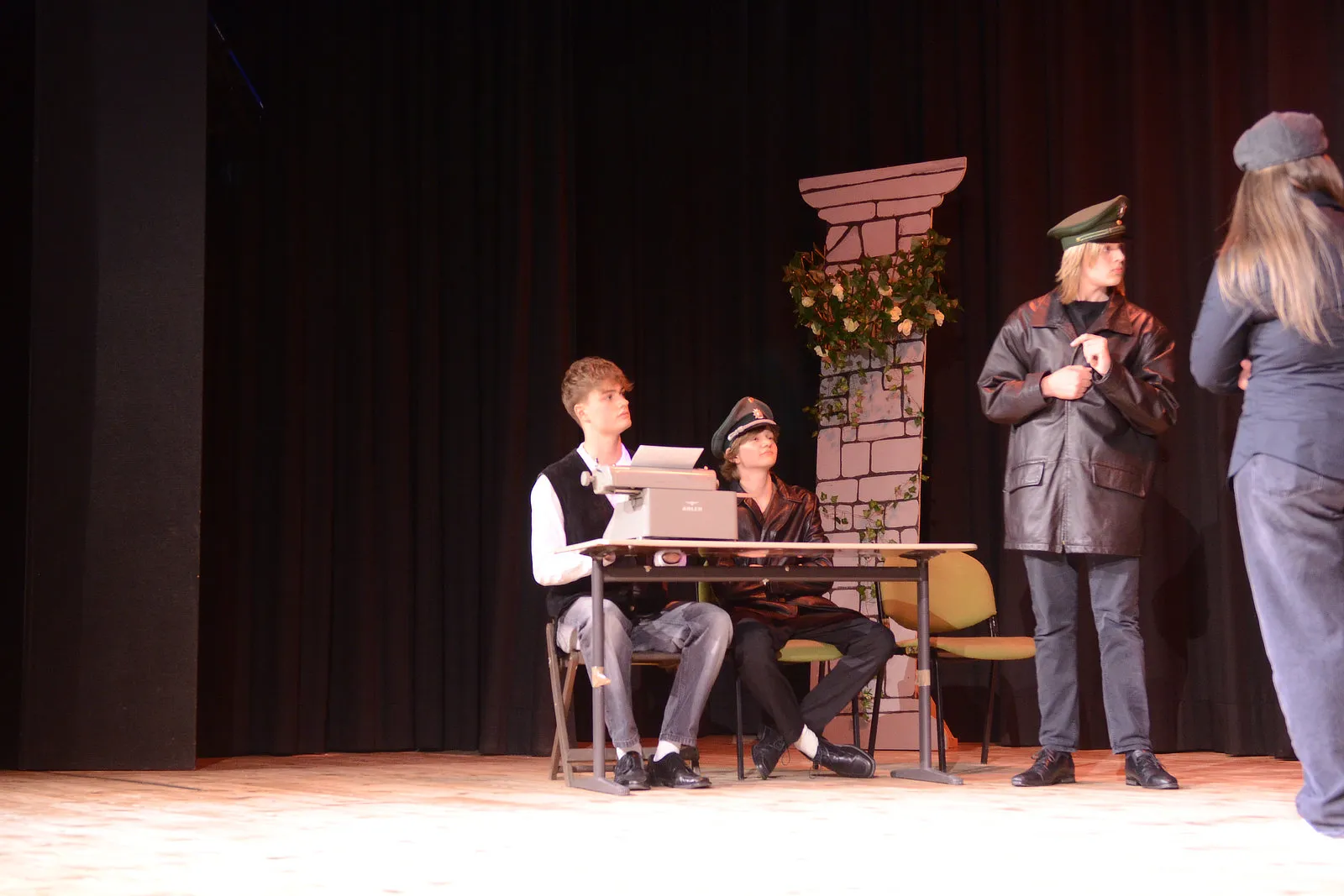 Szene aus der Theateraufführung „Viel Lärm um nichts“ mit drei Darstellern auf einer Bühne. Ein junger Mann tippt auf einer Schreibmaschine, während zwei weitere Personen im Hintergrund stehen.