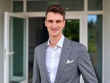 Ein Mann mit kurzen braunen Haaren lächelt in die Kamera. Er trägt einen grauen Anzug mit einem weißen Hemd und eine weiße Taschentuch in der Brusttasche. Im Hintergrund ist eine offene Tür zu sehen.