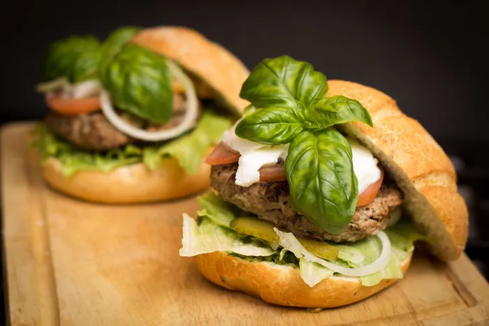 Zwei Burger mit Fleischpatty, Salat, Tomate, Zwiebeln und großen Basilikumblättern auf einem Holzbrett.