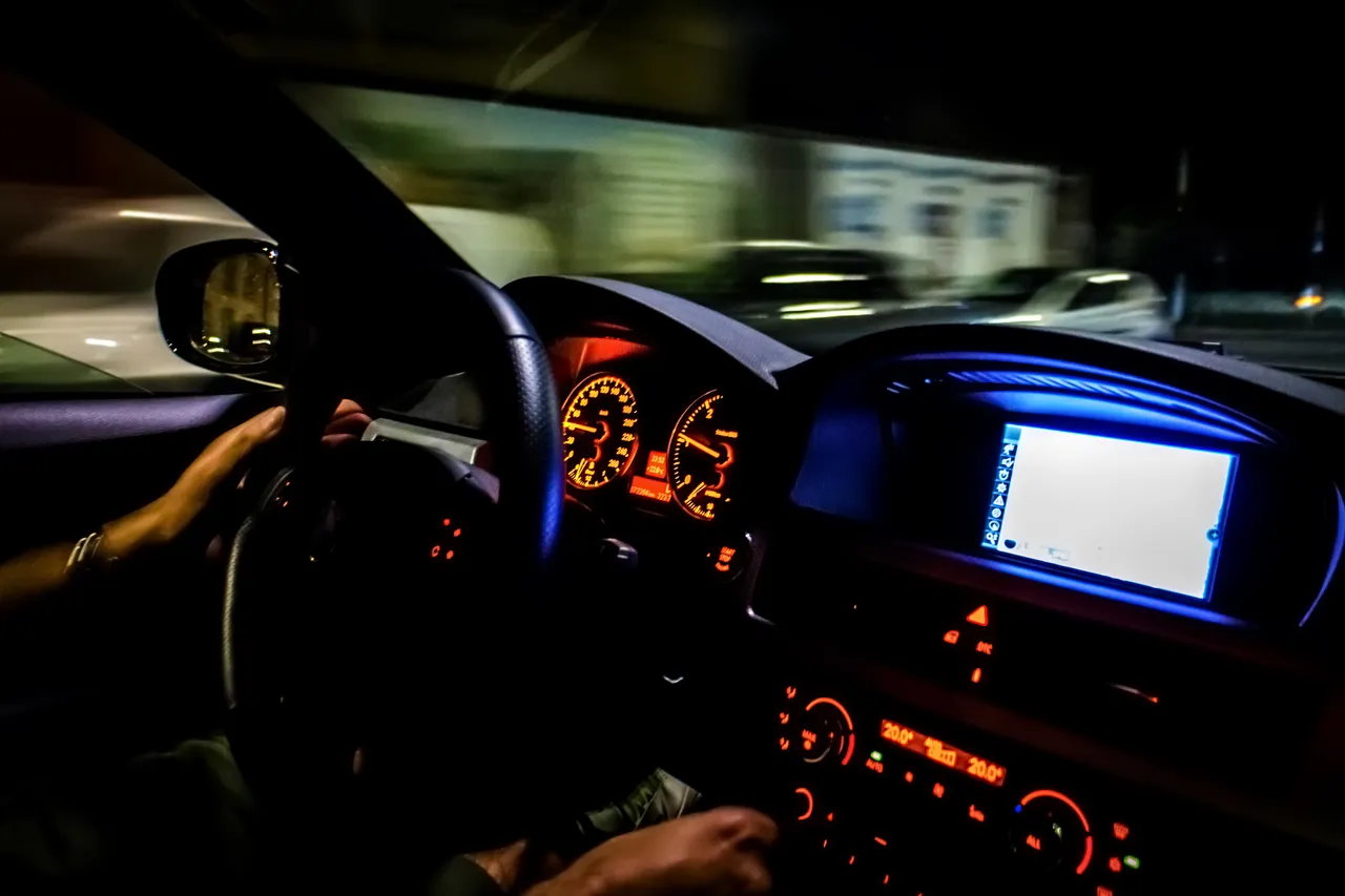 Innenansicht eines Autos bei Nacht mit beleuchtetem Armaturenbrett und Händen am Lenkrad vor einer durch Bewegungsunschärfe verschwommenen Umgebung.