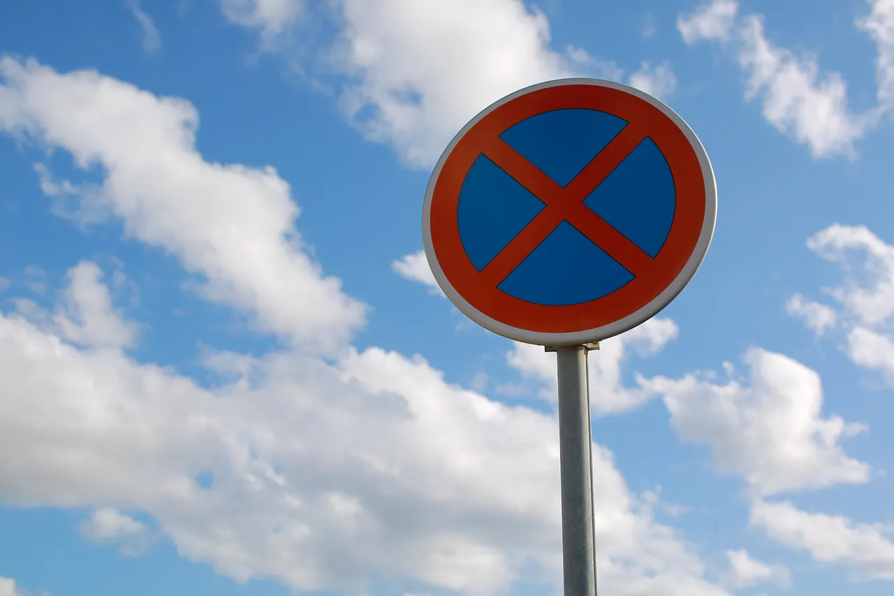 Rundes Verkehrszeichen mit blauem Hintergrund, rotem Rand und einem roten Kreuz vor einem blau bewölkten Himmel.