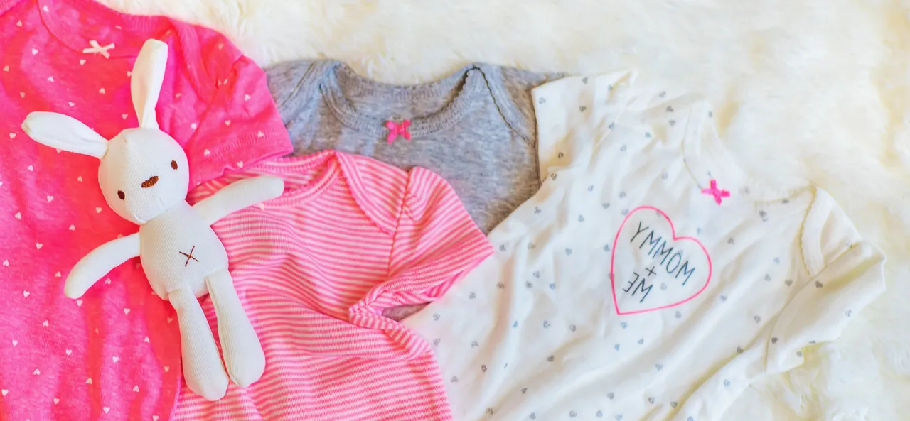Mehrere Babykleidungsstücke und ein Stofftier liegen auf einem weißen, flauschigen Untergrund. Die Kleidung umfasst rosa und graue Oberteile mit Herz- und Streifenmustern.