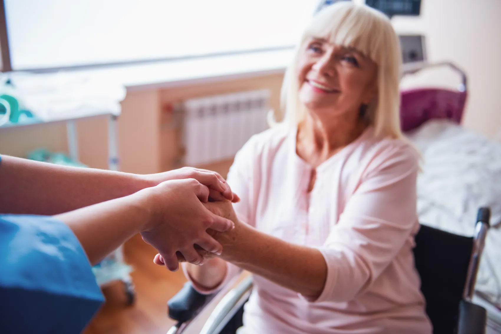 Eine ältere Frau mit blondem Haar lächelt, während sie die Hand einer Person in einem blauen Pflegehemd hält. Im Hintergrund ist ein Krankenhausbett zu sehen.