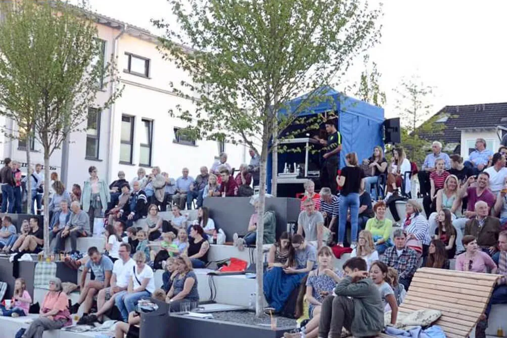Zuschauer sitzen und stehen vor einer Bühne unter einem blauen Zelt, umgeben von Bäumen und Häusern.