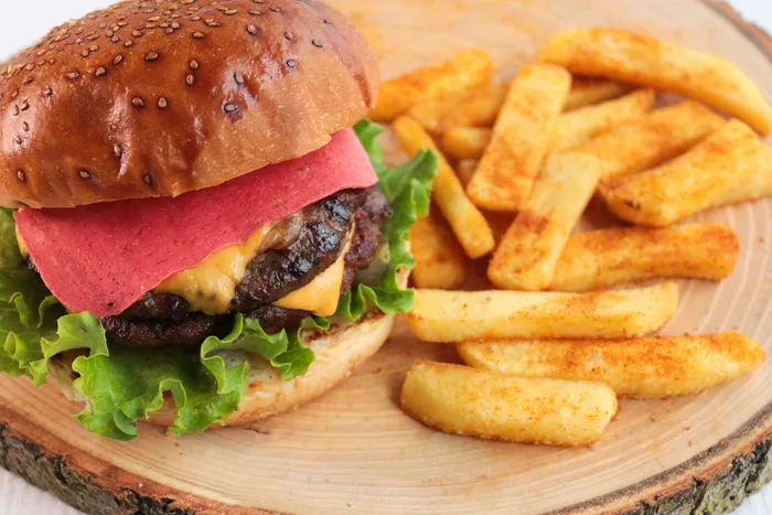 Burger mit Sesambrötchen, einer rosa Scheibe, Fleisch, Käse und Salat neben einer Portion Pommes Frites auf einem Holzbrett.