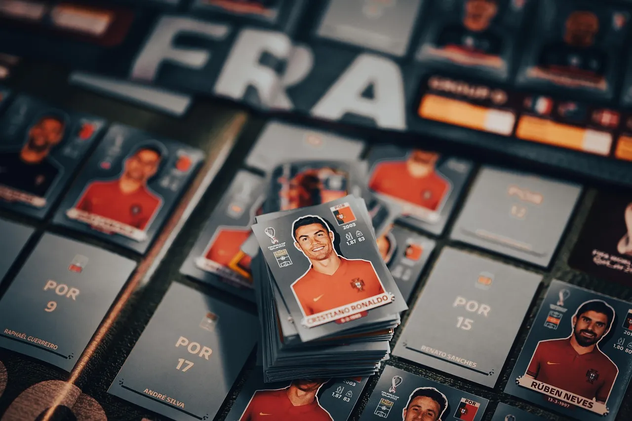 Sammlung portugiesischer Panini-Fußballspieler-Sammelkarten, darunter mehrere Karten von Cristiano Ronaldo, die auf einem Tisch ausgelegt sind. Im Hintergrund ist ein Schild mit der Aufschrift „FRA“ zu sehen.