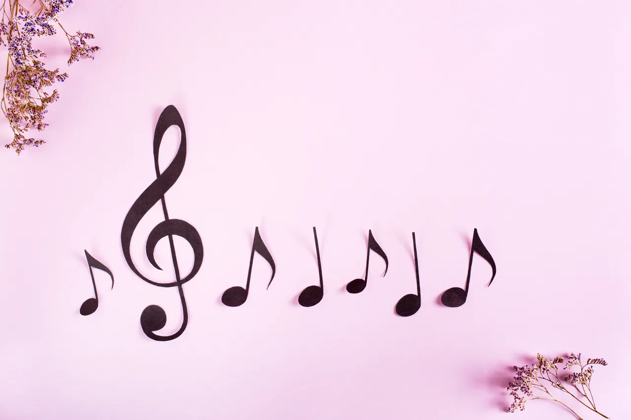 Schwarze Papiernoten und ein Violinschlüssel liegen auf einem hellrosa Hintergrund. Trockenblumen ergänzen das Arrangement.
