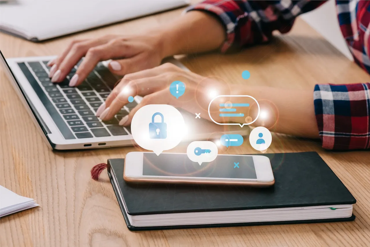 Hände tippen auf Laptop, darüber und neben einem Smartphone Icons für Sicherheit, Nutzerkonto und Schlüssel.