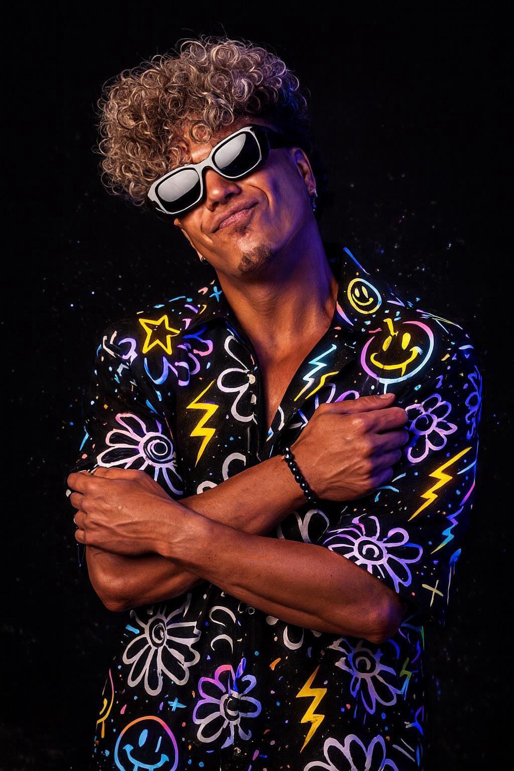 DJ Laky mit lockigem Haar und Sonnenbrille umarmt sich selbst, bekleidet mit einem Hemd mit bunten Kritzeleien vor schwarzem Hintergrund.