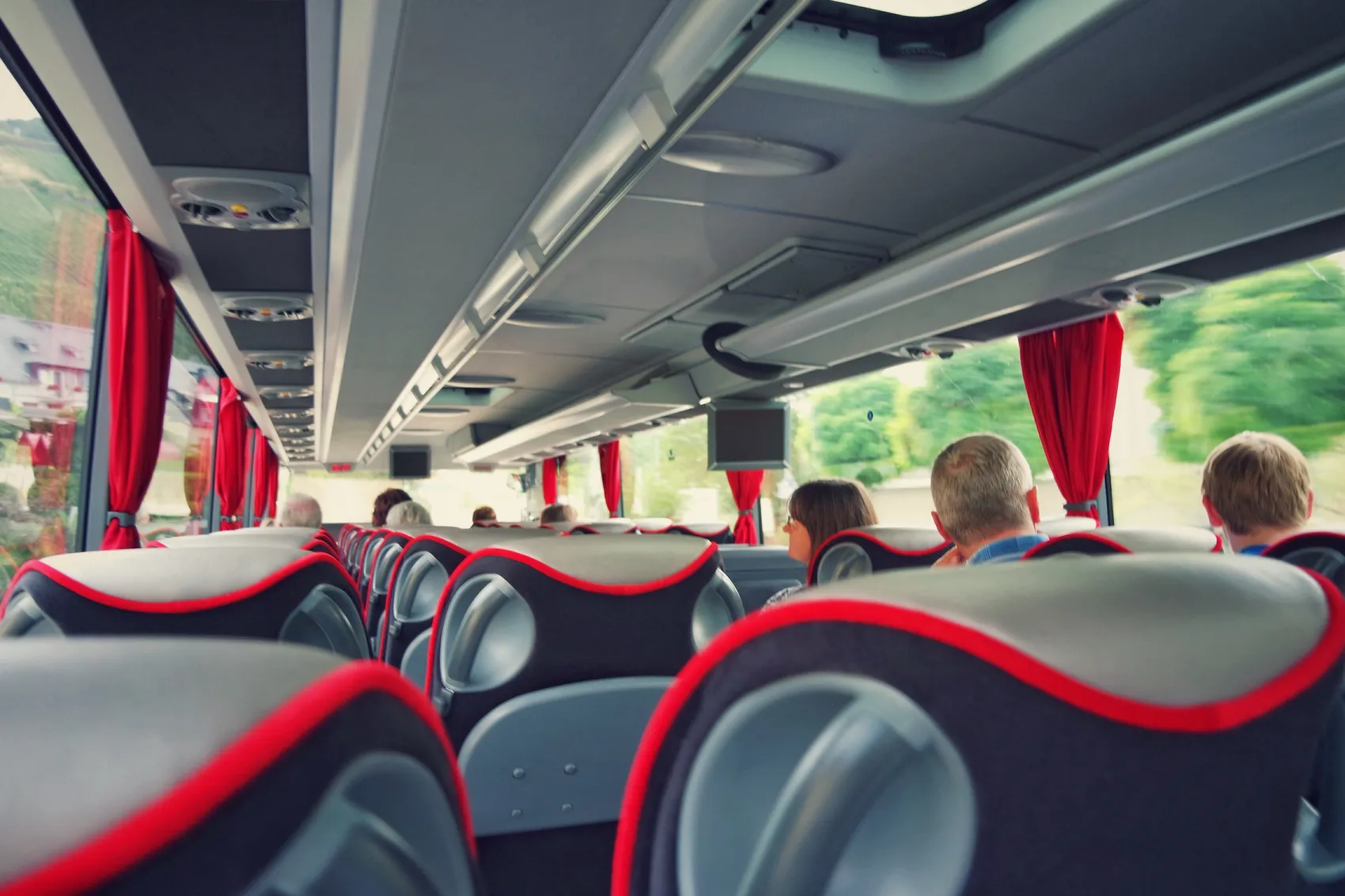 Reisende sitzen in einem Bus mit roten Vorhängen und schauen aus dem Fenster in die Landschaft. Der Innenraum ist hell erleuchtet.