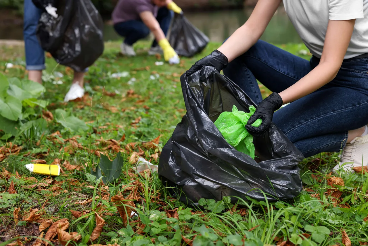 Eine Person mit schwarzen Handschuhen sammelt Müll in einem schwarzen Plastiksack auf einer Wiese ein.