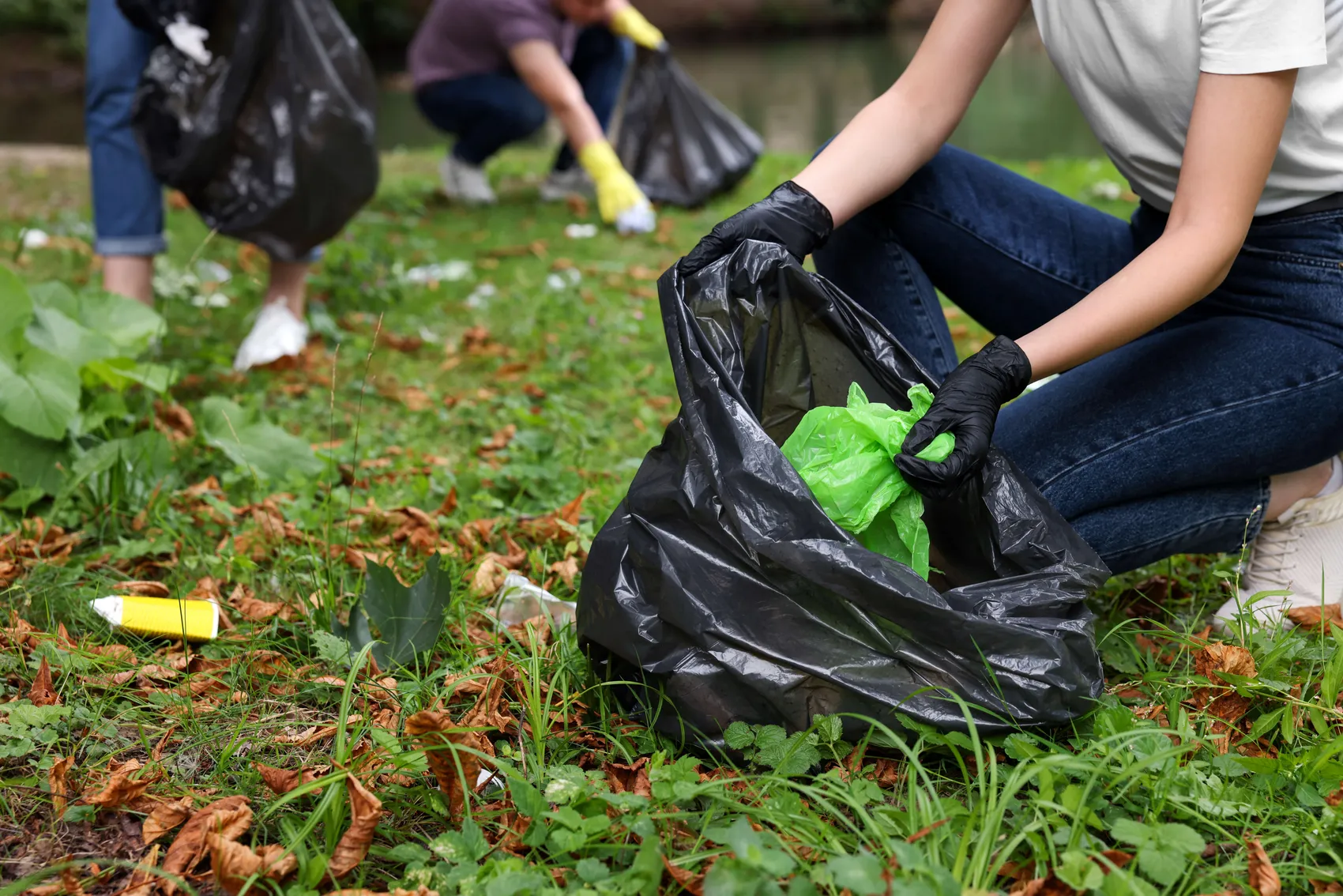 Eine Person mit schwarzen Handschuhen sammelt Müll in einem schwarzen Plastiksack auf einer Wiese ein.