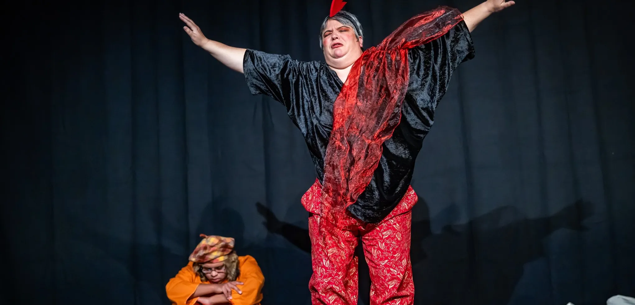 Darstellerin mit hoch erhobenen Armen und rotem Tuch auf der Bühne, im Hintergrund eine weitere Person mit Kopftuch.