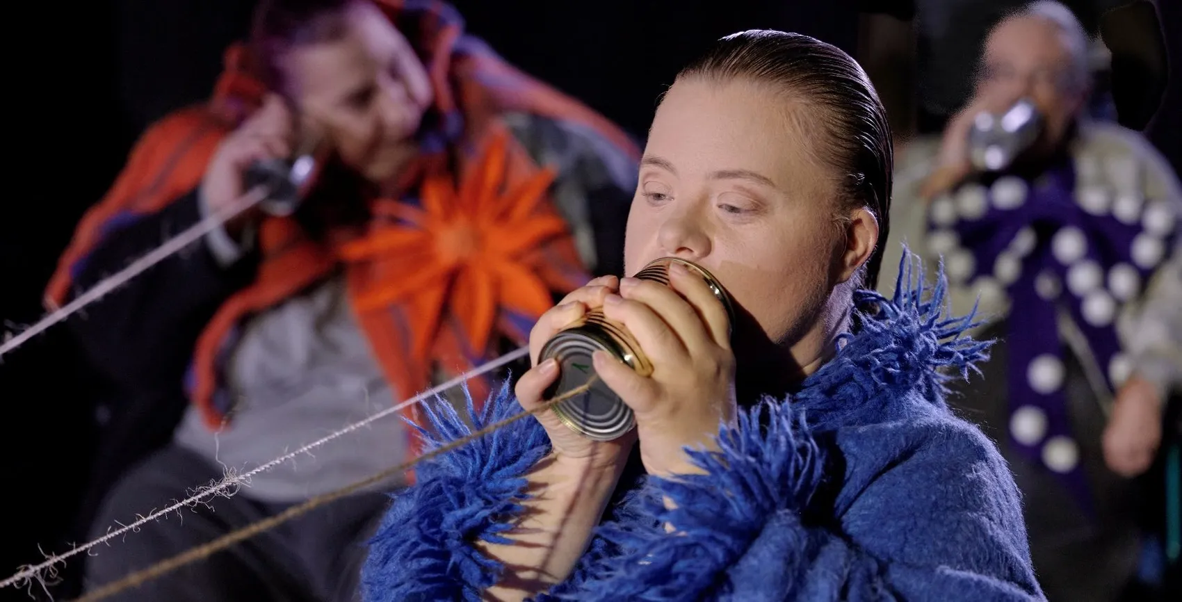 Ein Mann mit zurückgegeltem Haar spricht in eine Konservendose, die an einer Schnur befestigt ist. Im Hintergrund sind weitere Personen mit ähnlichen Konservendosen zu sehen.