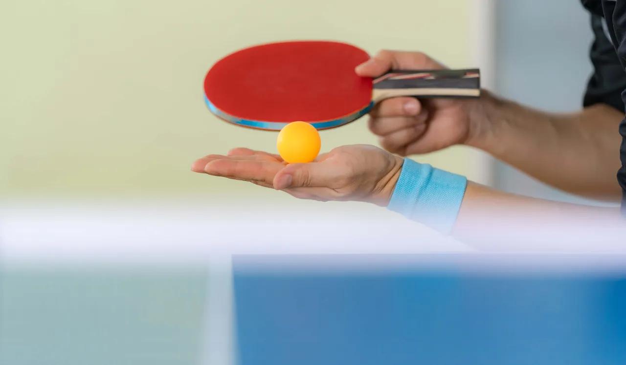 Eine Hand hält einen gelben Tischtennisball, die andere einen roten Tischtennisschläger.