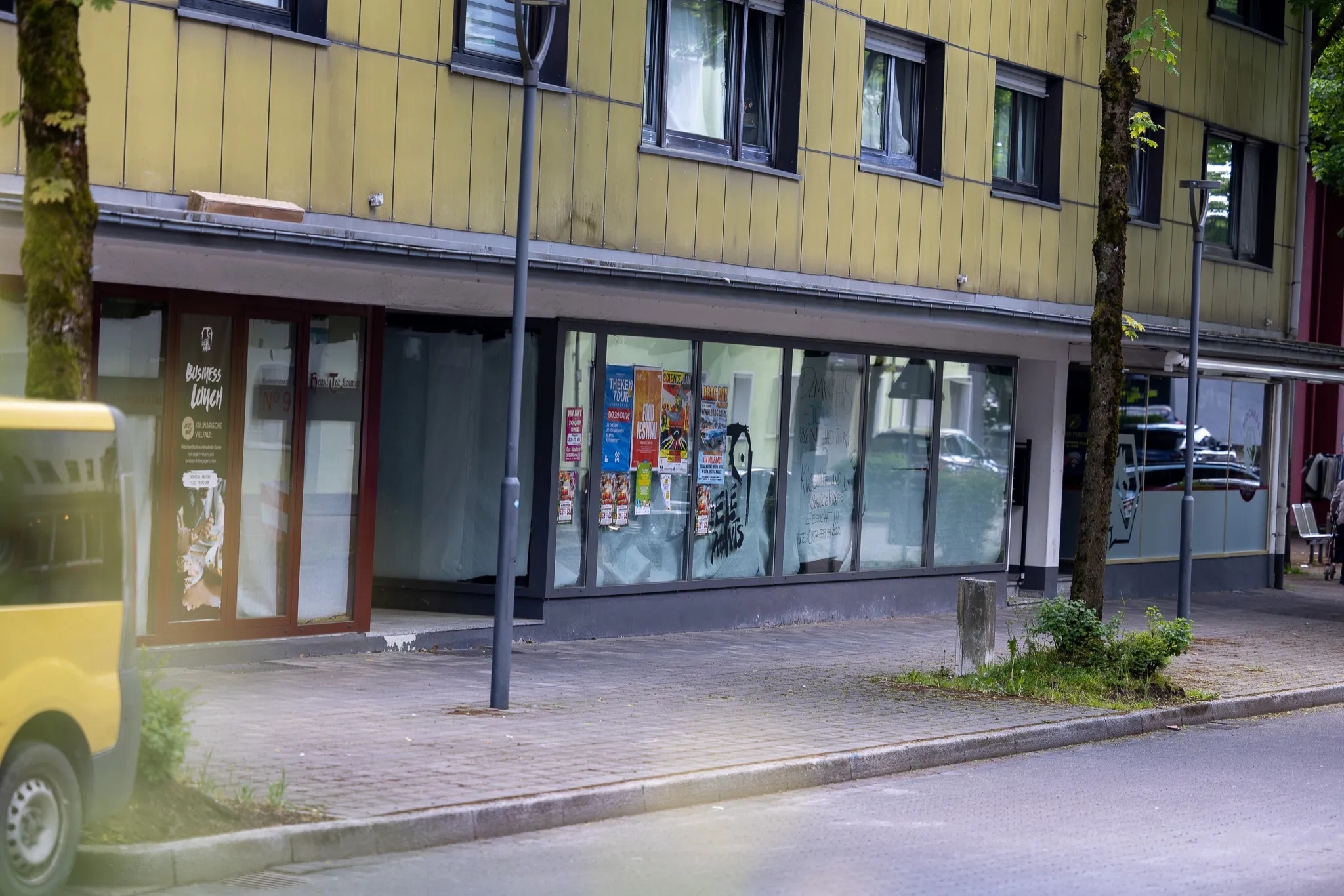 Gelbes Gebäude mit Schaufenstern und einem Eingangsbereich, an dem ein Plakat für einen Business Lunch hängt. Vor dem Gebäude befindet sich ein Gehweg und eine Straße.