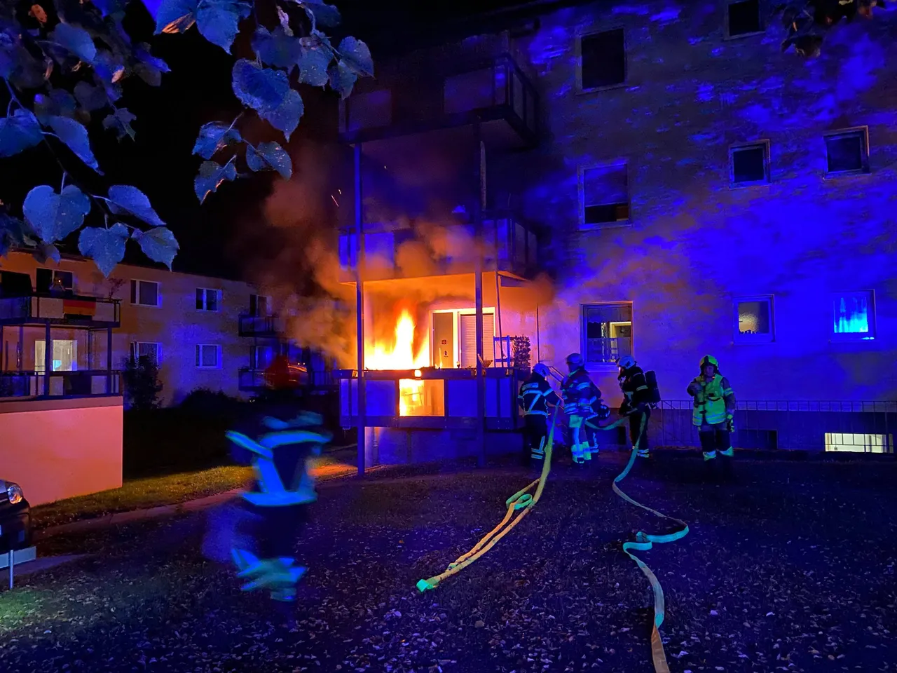 Feuerwehrleute im Einsatz bei einem nächtlichen Gebäudebrand, bei dem Flammen aus einem Balkon schlagen.