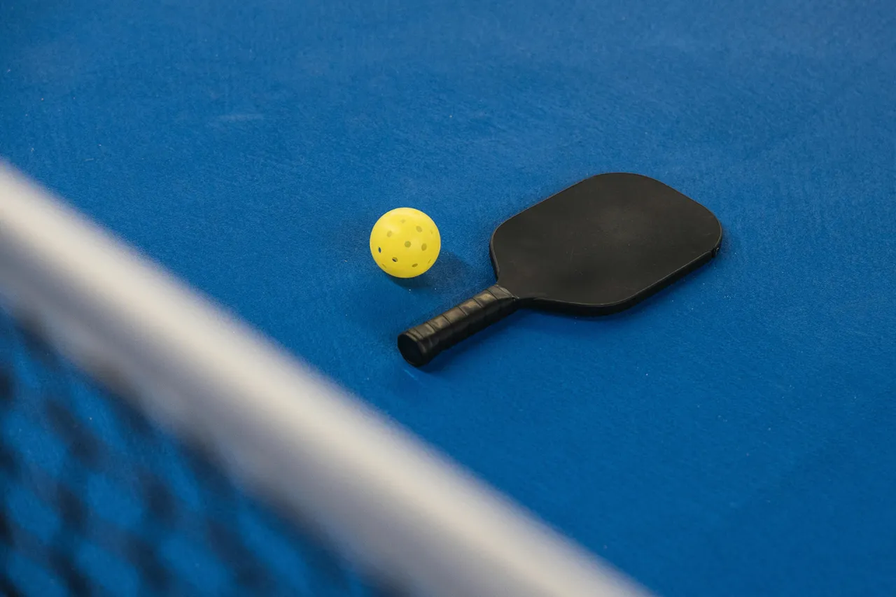 Gelber Pickleball und schwarzer Schläger auf blauem Spielfeld, Netz im Vordergrund.